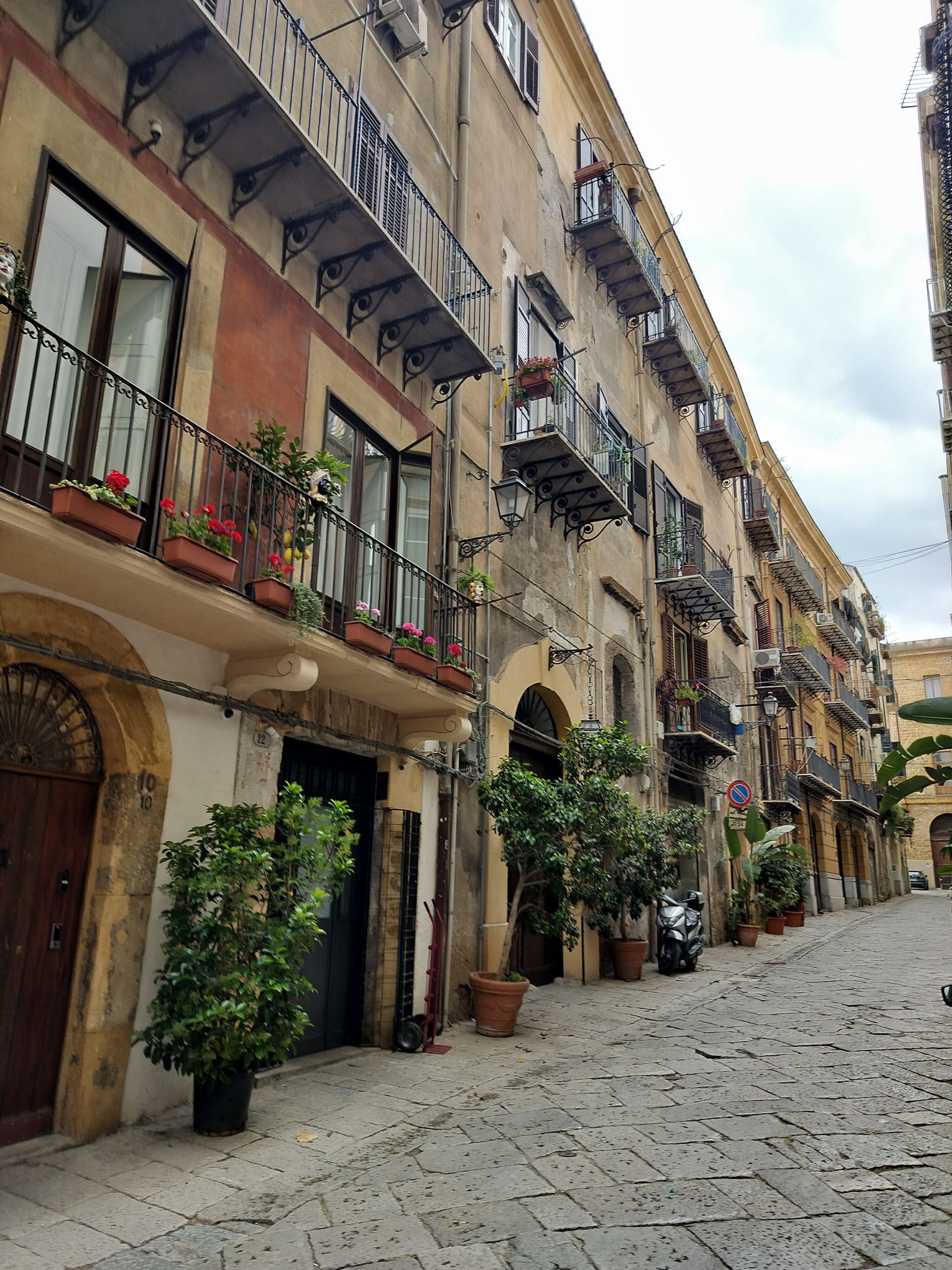 A narrow cobblestone street in a European city, lined with old buildings with balconies and potted plants.