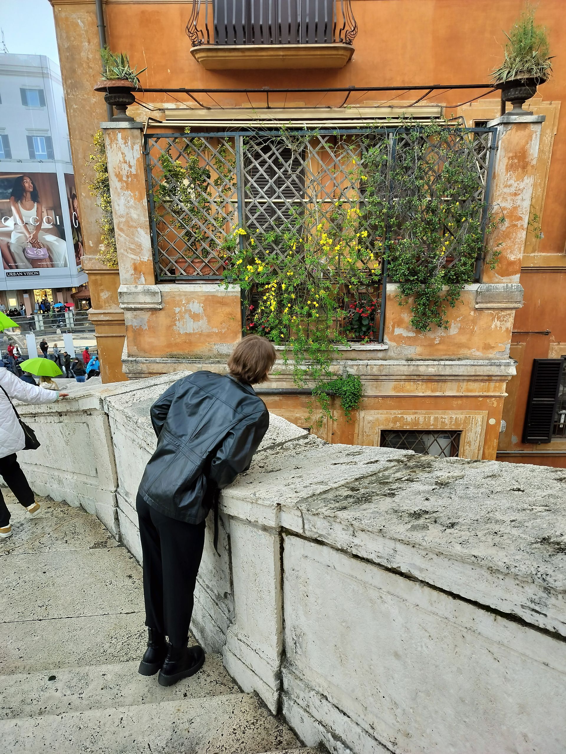 Person looking over a stone wall at a weathered building with a window covered in greenery.