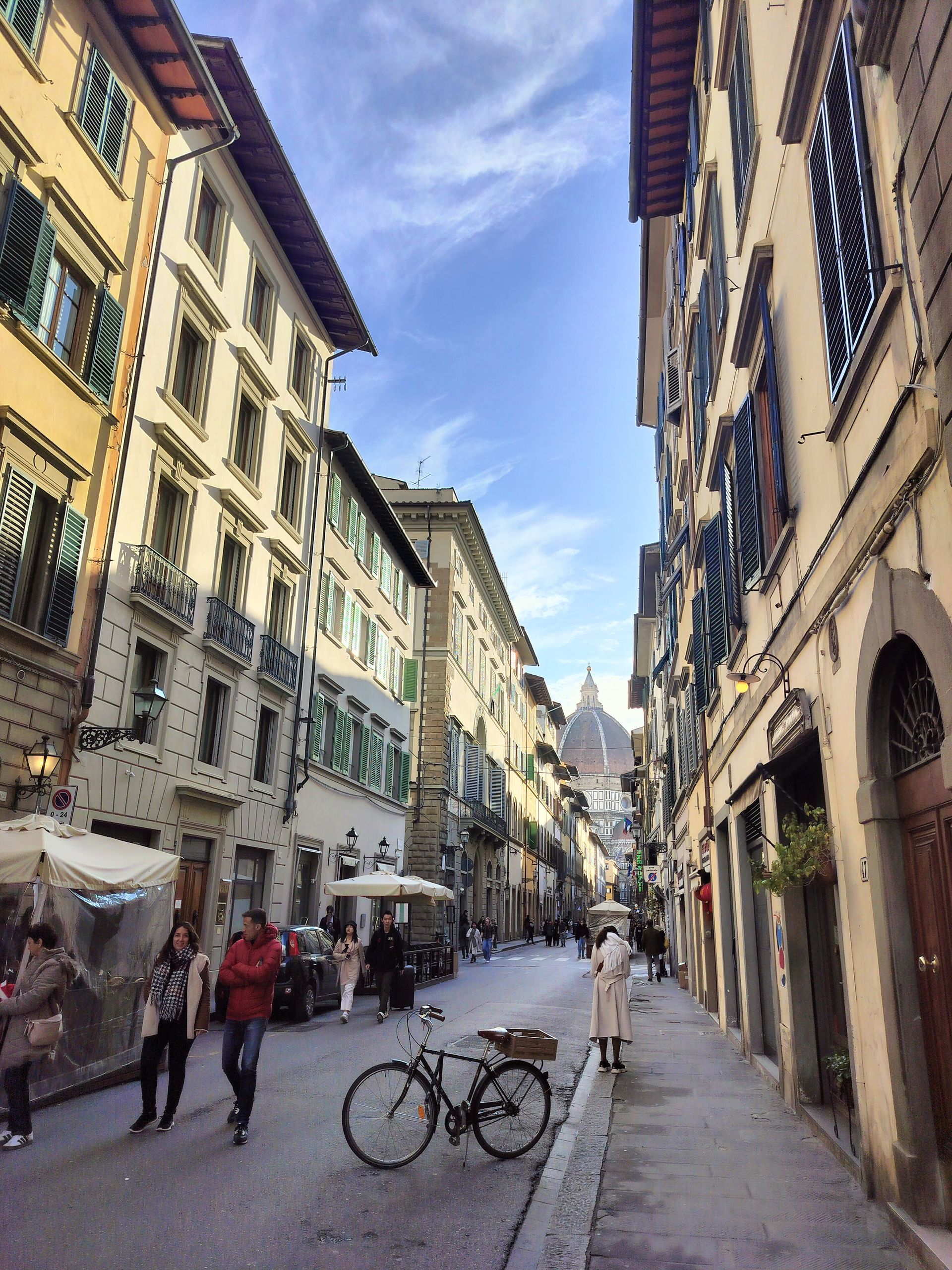 Narrow street in Florence, Italy, with historic buildings, a bicycle, and pedestrians. Blue sky with dome in distance.