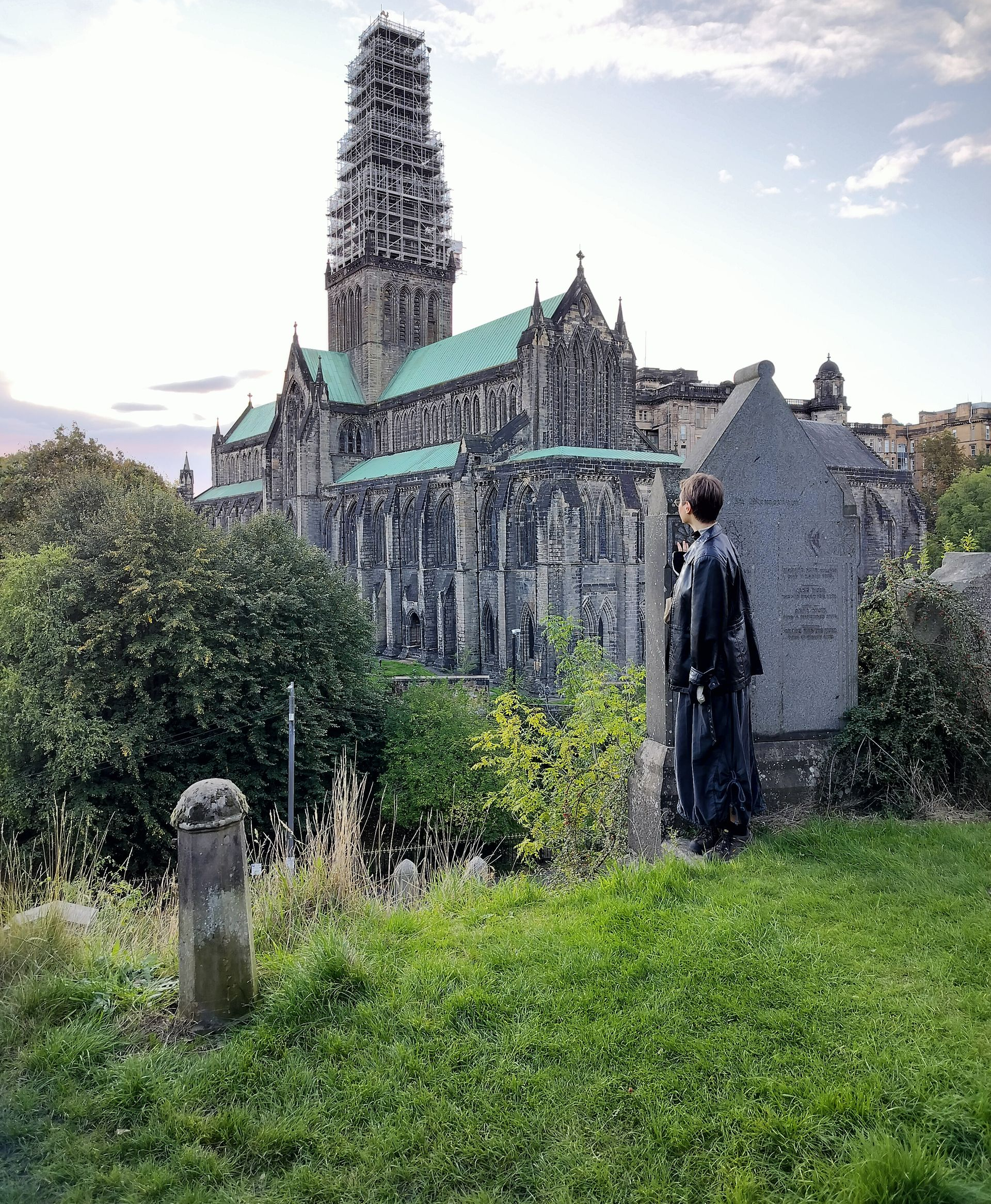 Person in black coat looking at a large cathedral. Stone building, green grass, and overcast sky.