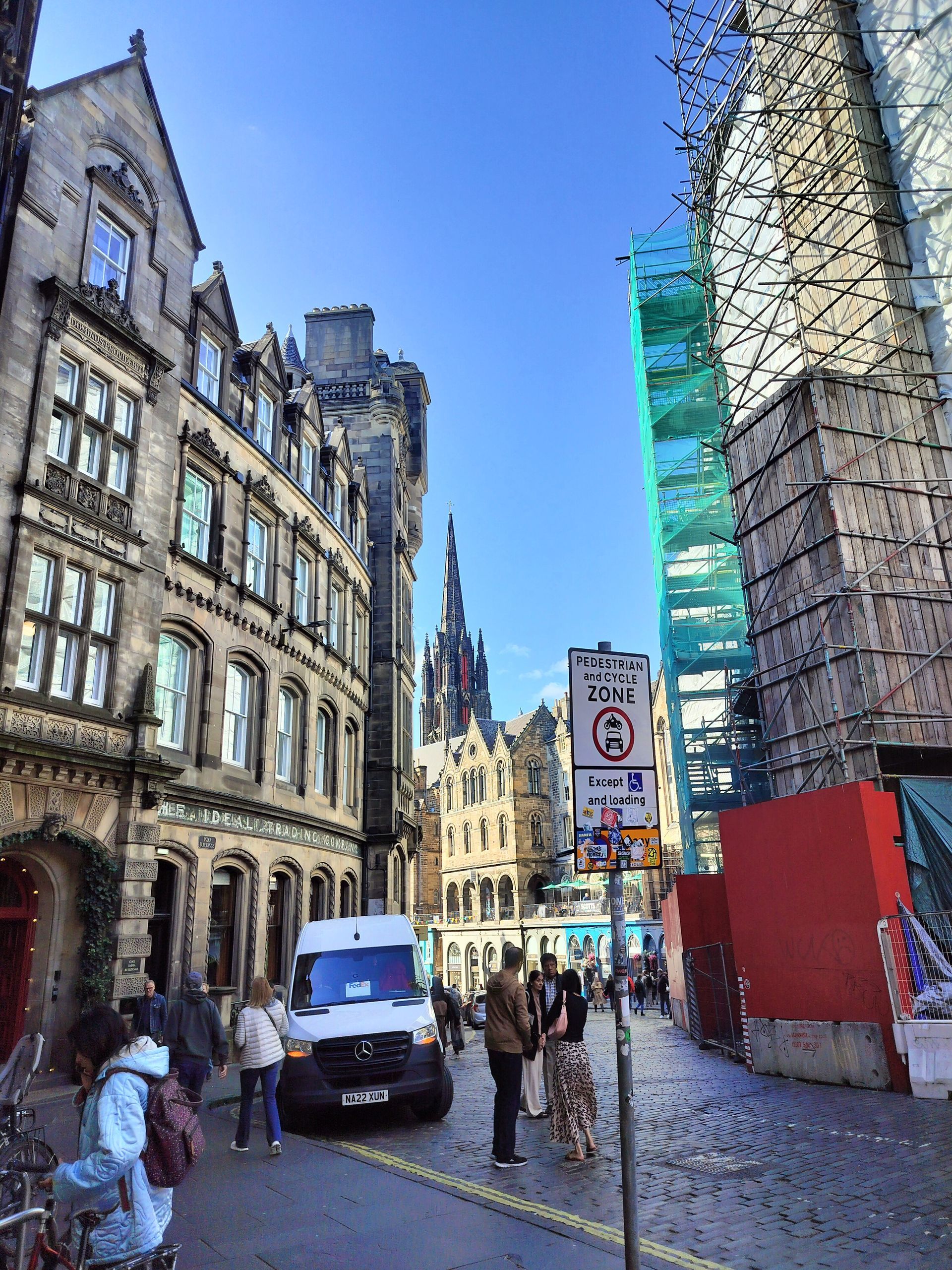 Cobblestone street scene in Edinburgh. Buildings line the street, a van and people are present. Blue sky overhead.