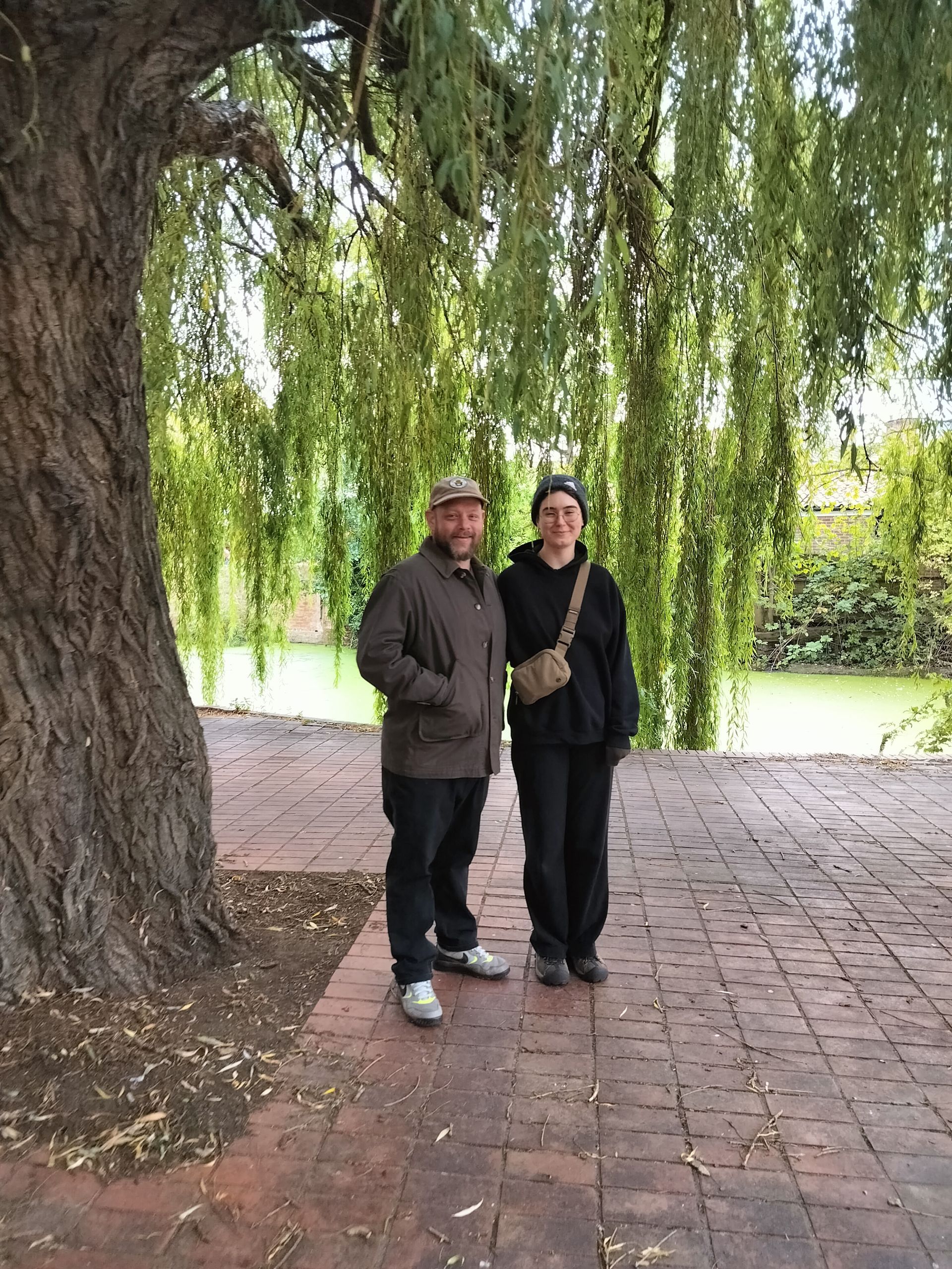 Two people standing beneath a weeping willow tree on a brick pathway, next to a body of water.