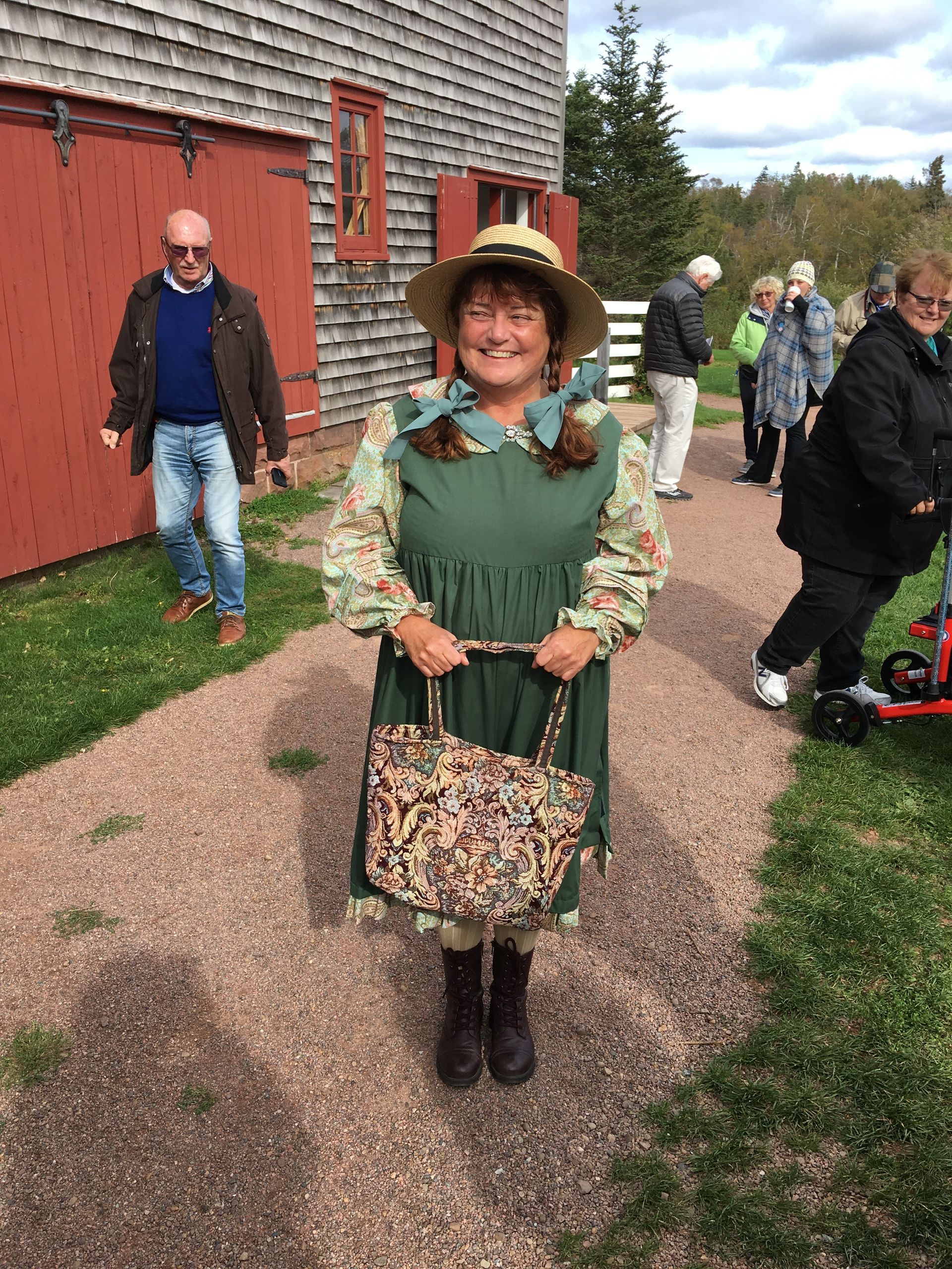 Woman in costume, holding a patterned bag, stands on a gravel path near a red barn; people in background.