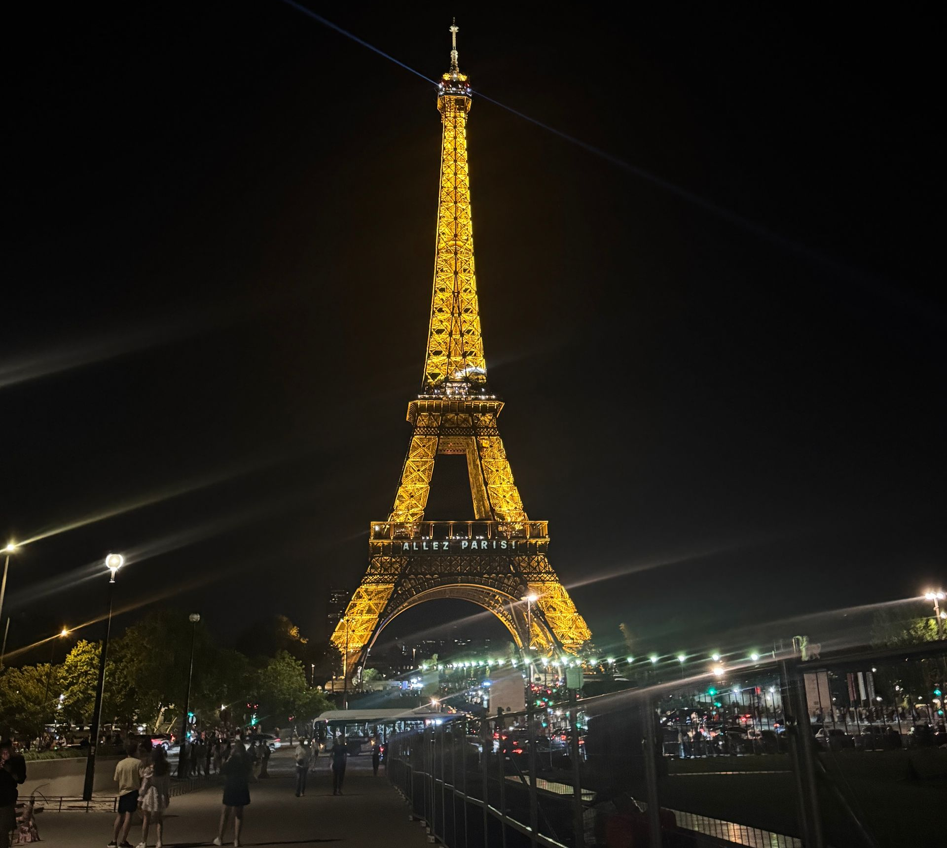 Eiffel Tower illuminated at night; people on the ground with some lights and the Seine River in the background.