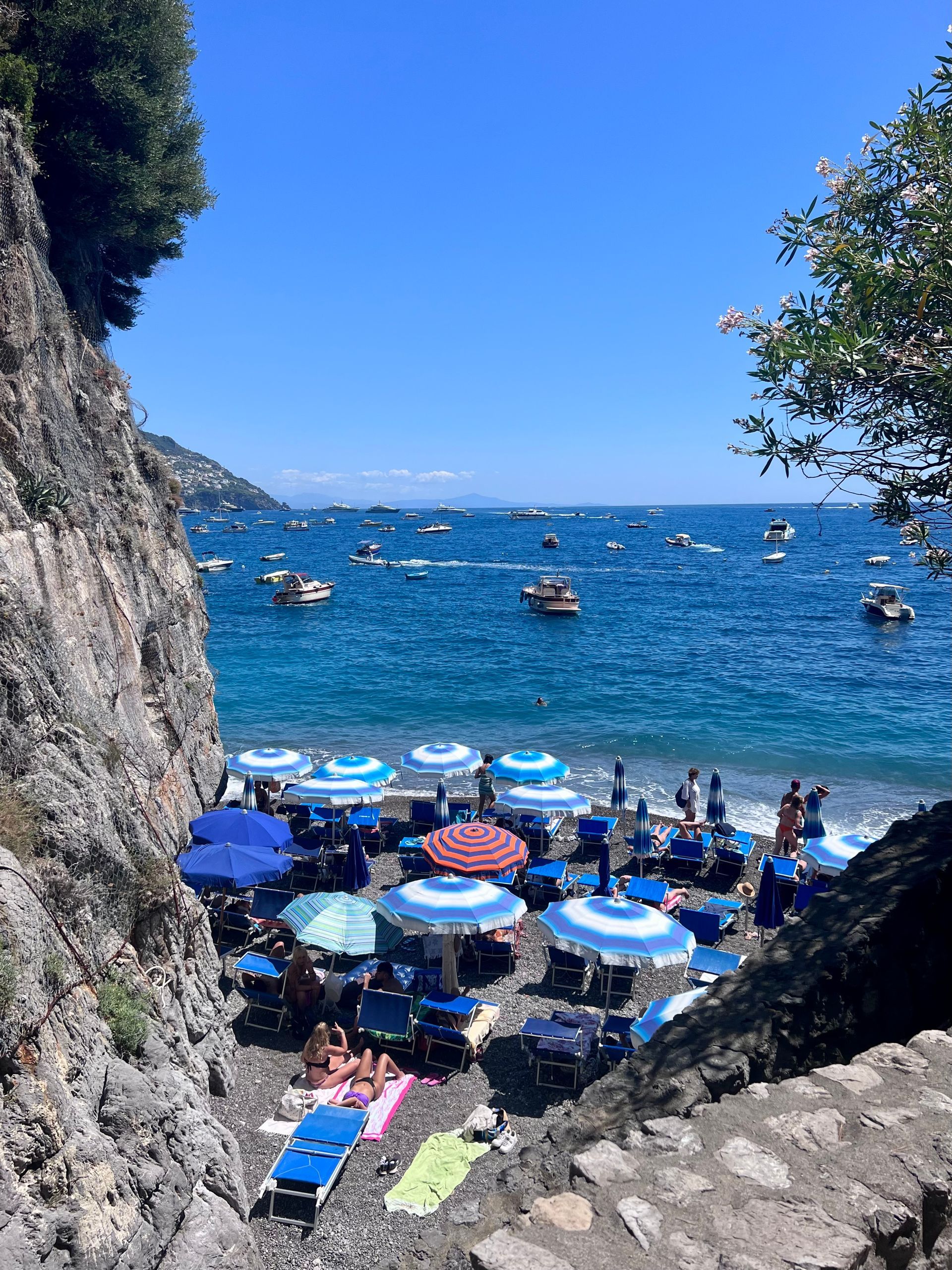 Beach with blue umbrellas and boats on the sea, framed by cliffs under a bright sky.