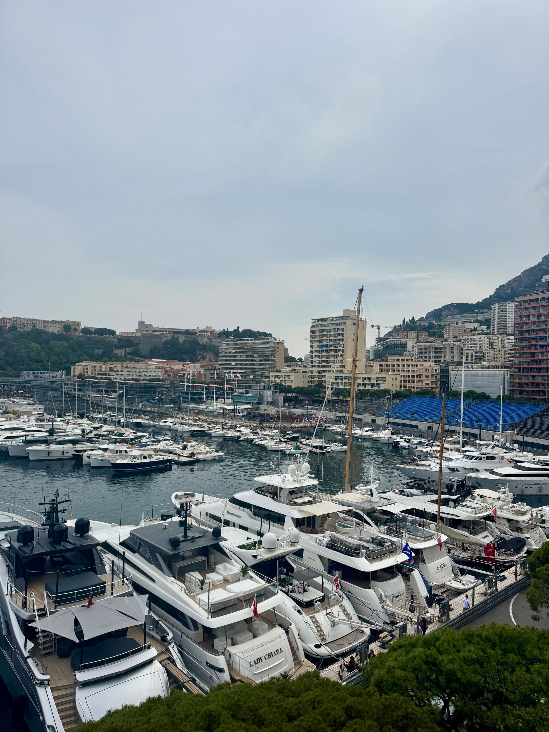 Yachts docked in a harbor, buildings on a hillside, cloudy sky.