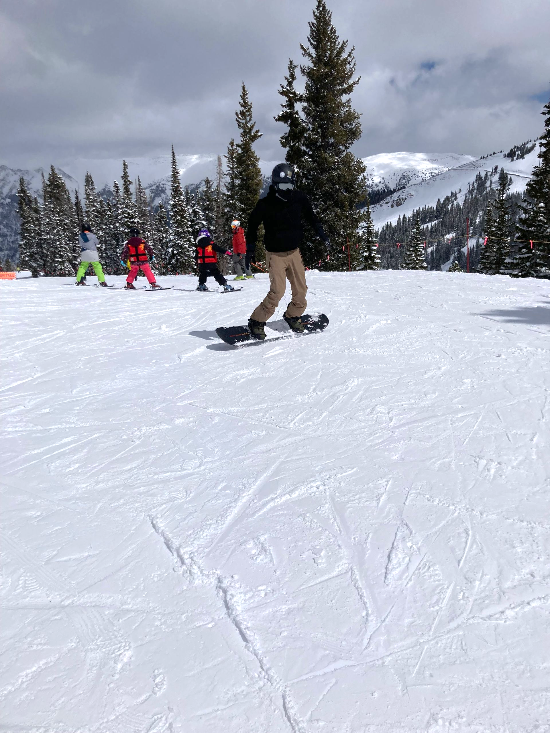 Person snowboarding down a snowy slope, arms outstretched. Other skiers and trees in the background.