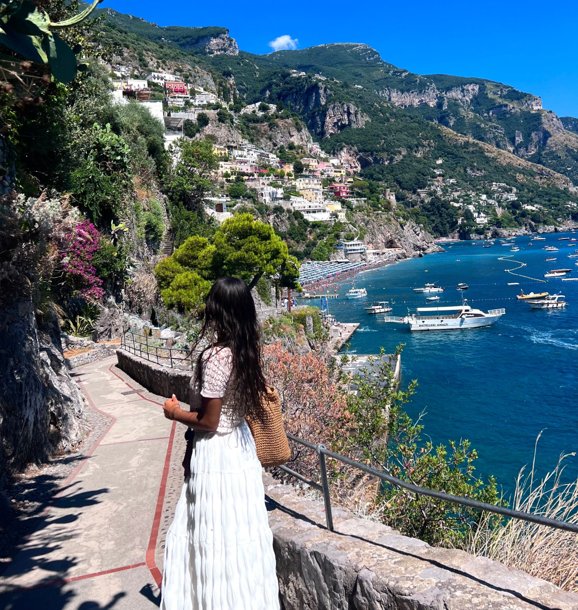 Woman overlooking Positano, Italy, coastline. Buildings dot hillside, boats in water, blue sky.