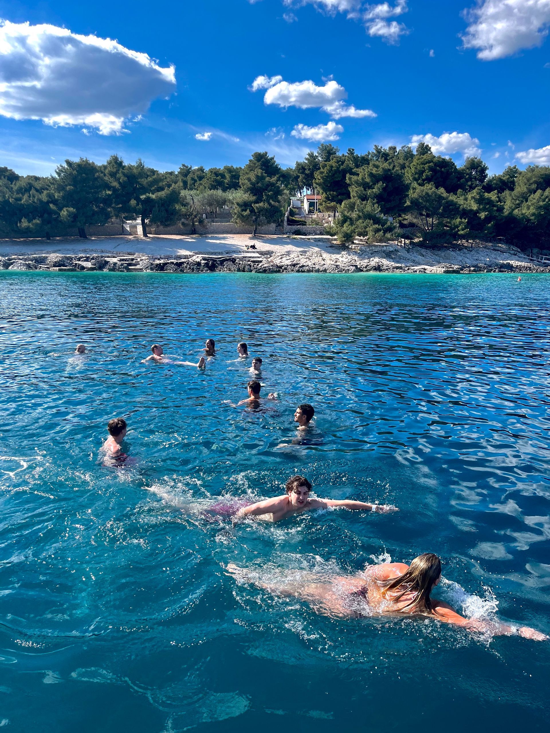 People swimming in turquoise water near a tree-lined shore under a blue sky with clouds.