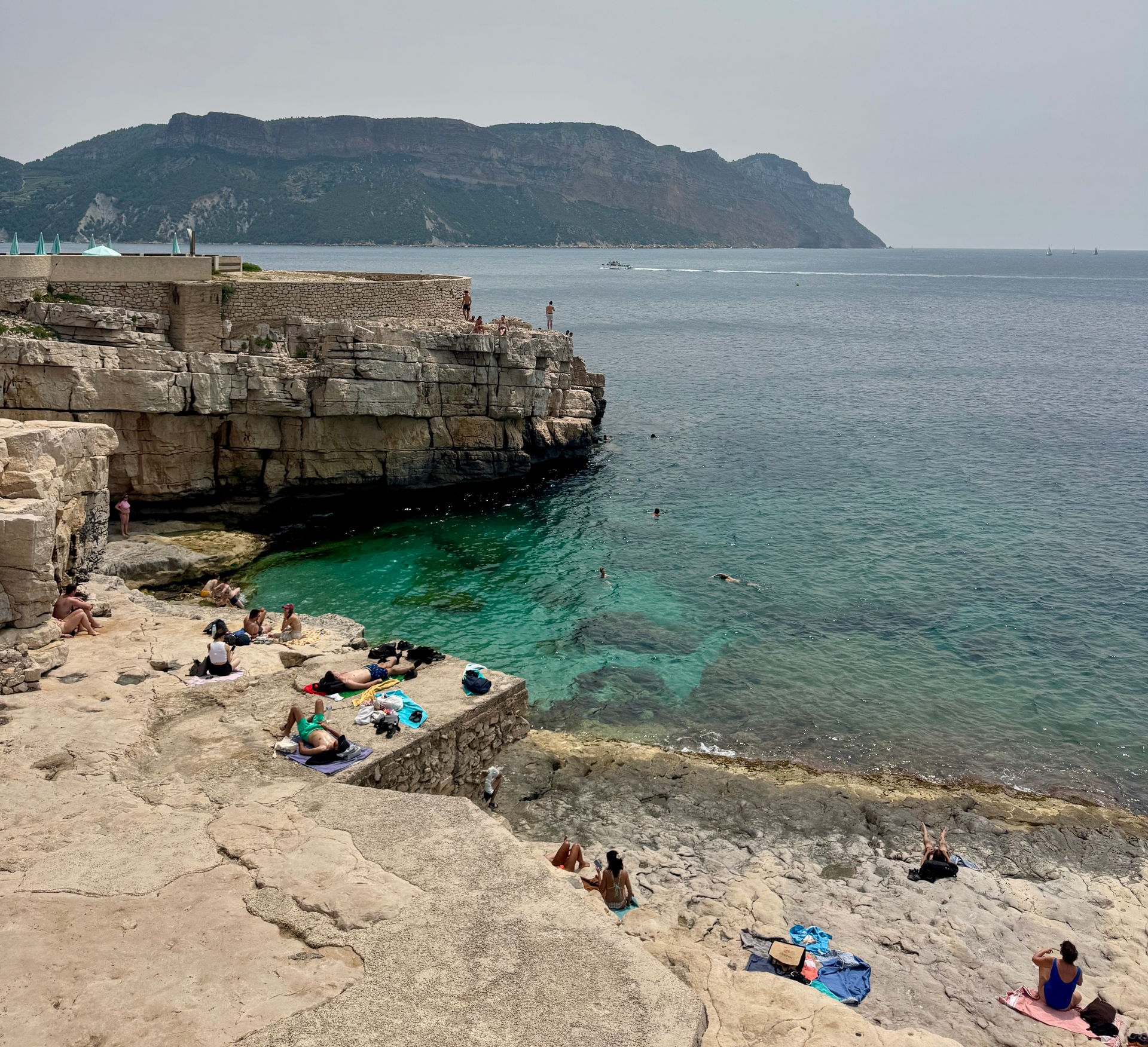 Rocky beach with clear turquoise water, people sunbathing. Mountains in the distance.
