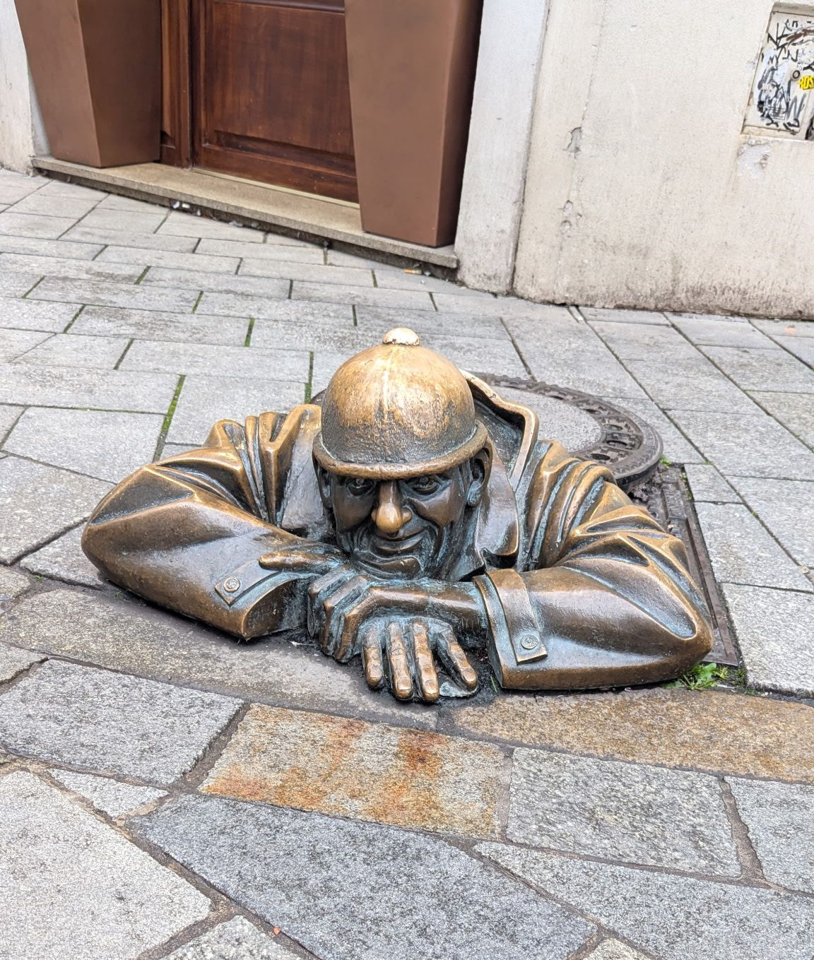 Bronze statue of a man peering out of a manhole in a city street.
