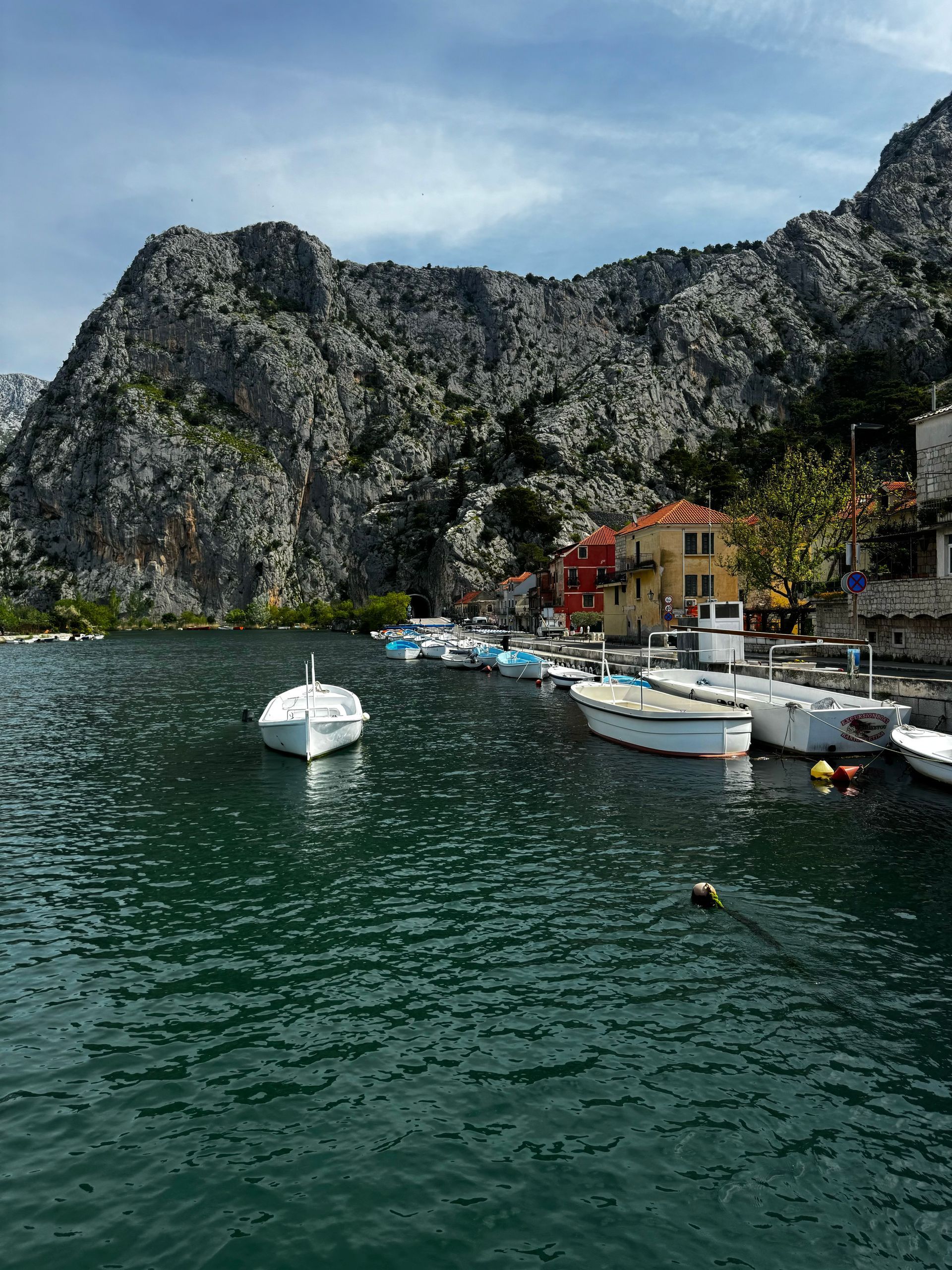 Boats docked along a waterfront, with a large rocky mountain in the background, under a partly cloudy sky.
