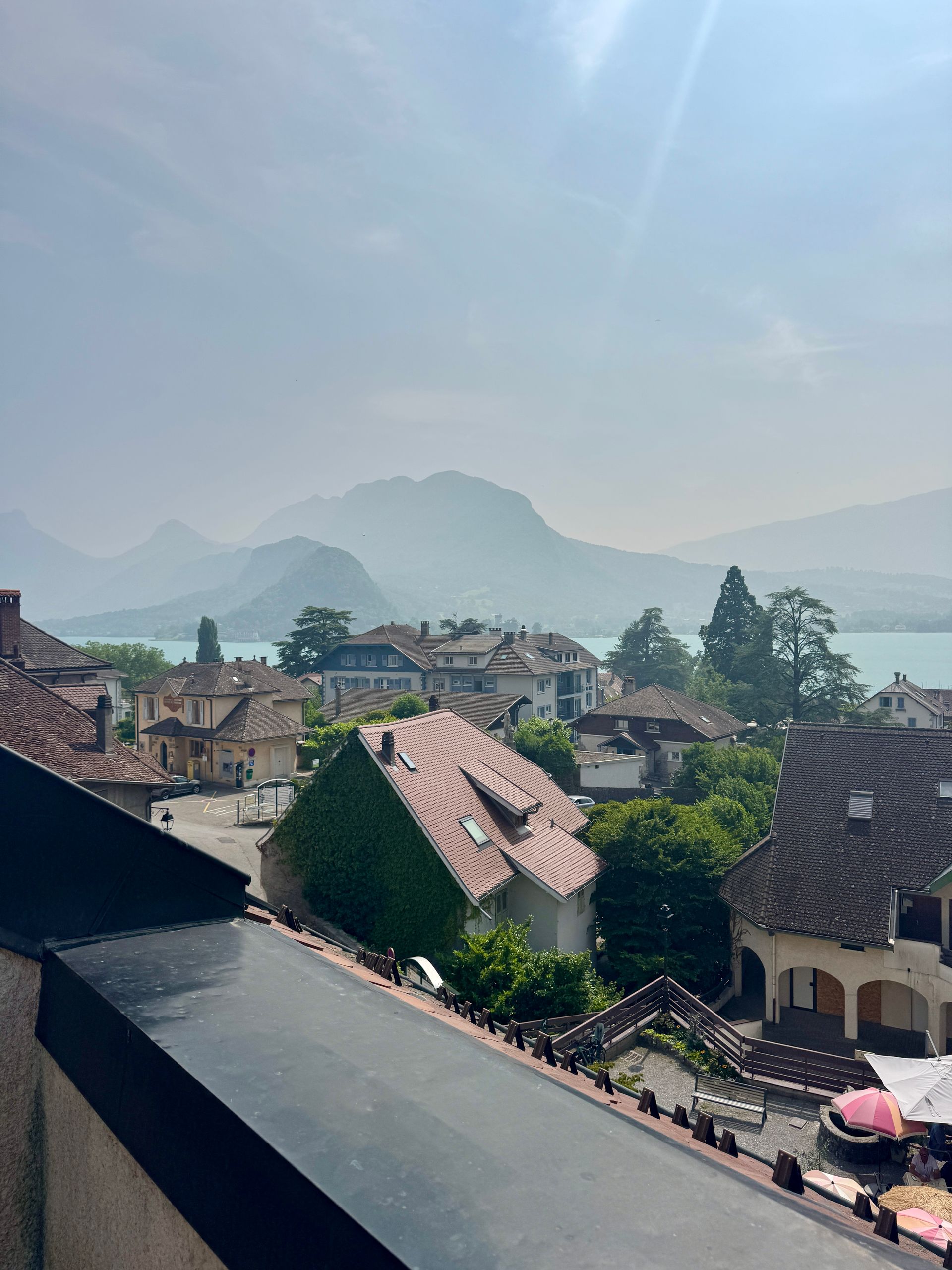 Overlooking European village with red-tiled roofs, lake, and mountains under a hazy sky.