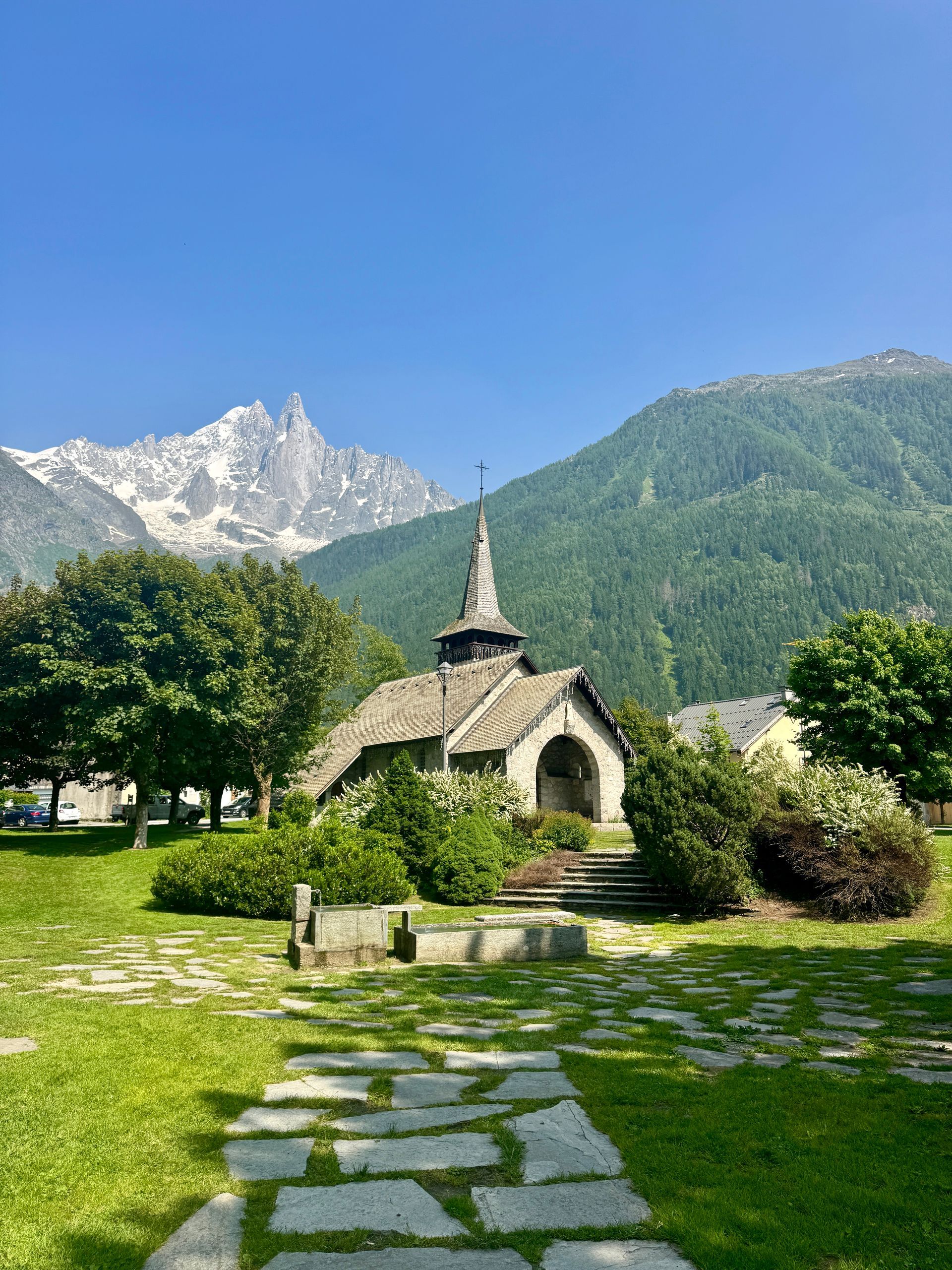 Stone chapel with steeple in front of mountains and green grass under a clear blue sky.