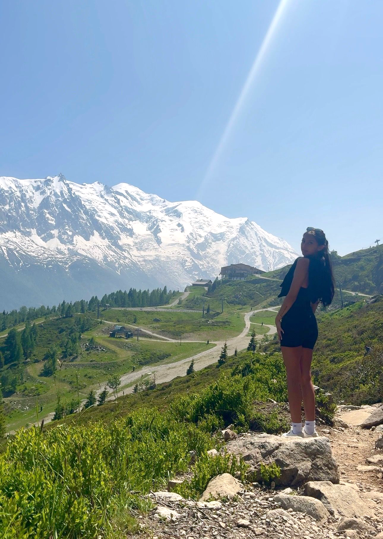 Woman stands on a mountain path, gazing up at the sun and snow-capped peaks. Sunny day.