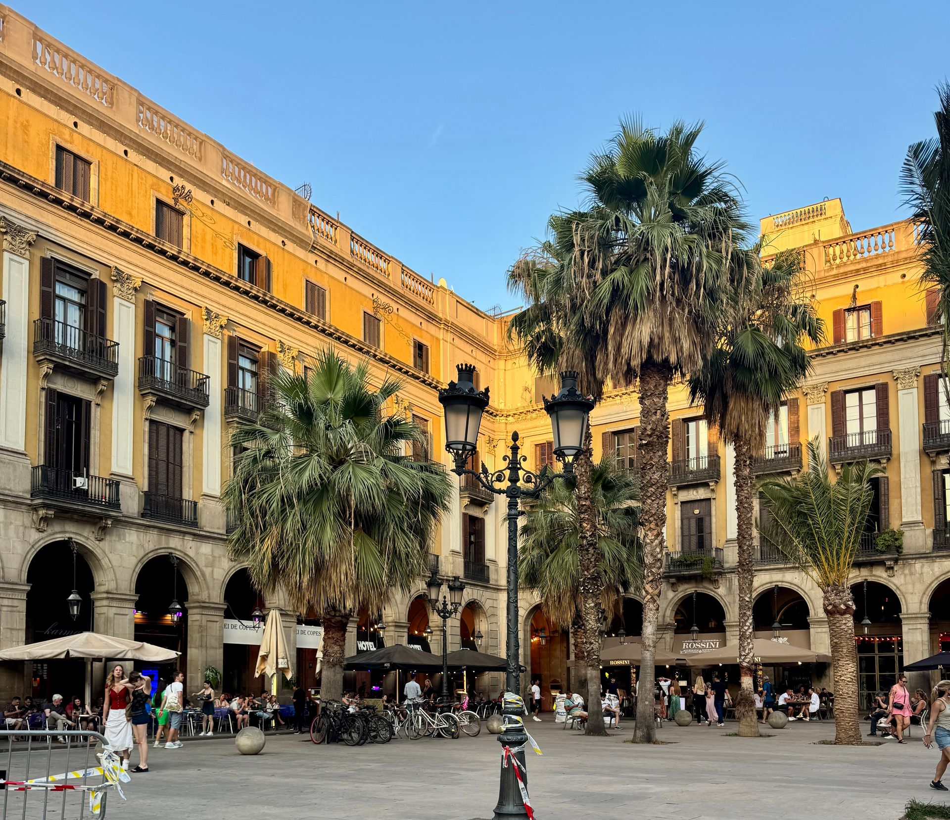 Square in Barcelona with arched buildings, palm trees, and outdoor seating; people gather.