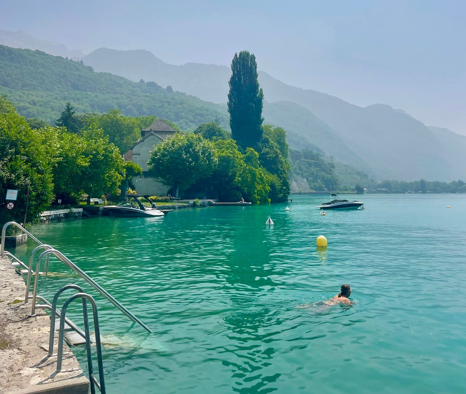 Lake with turquoise water, mountains, swimmer, boats, and trees on a sunny day.