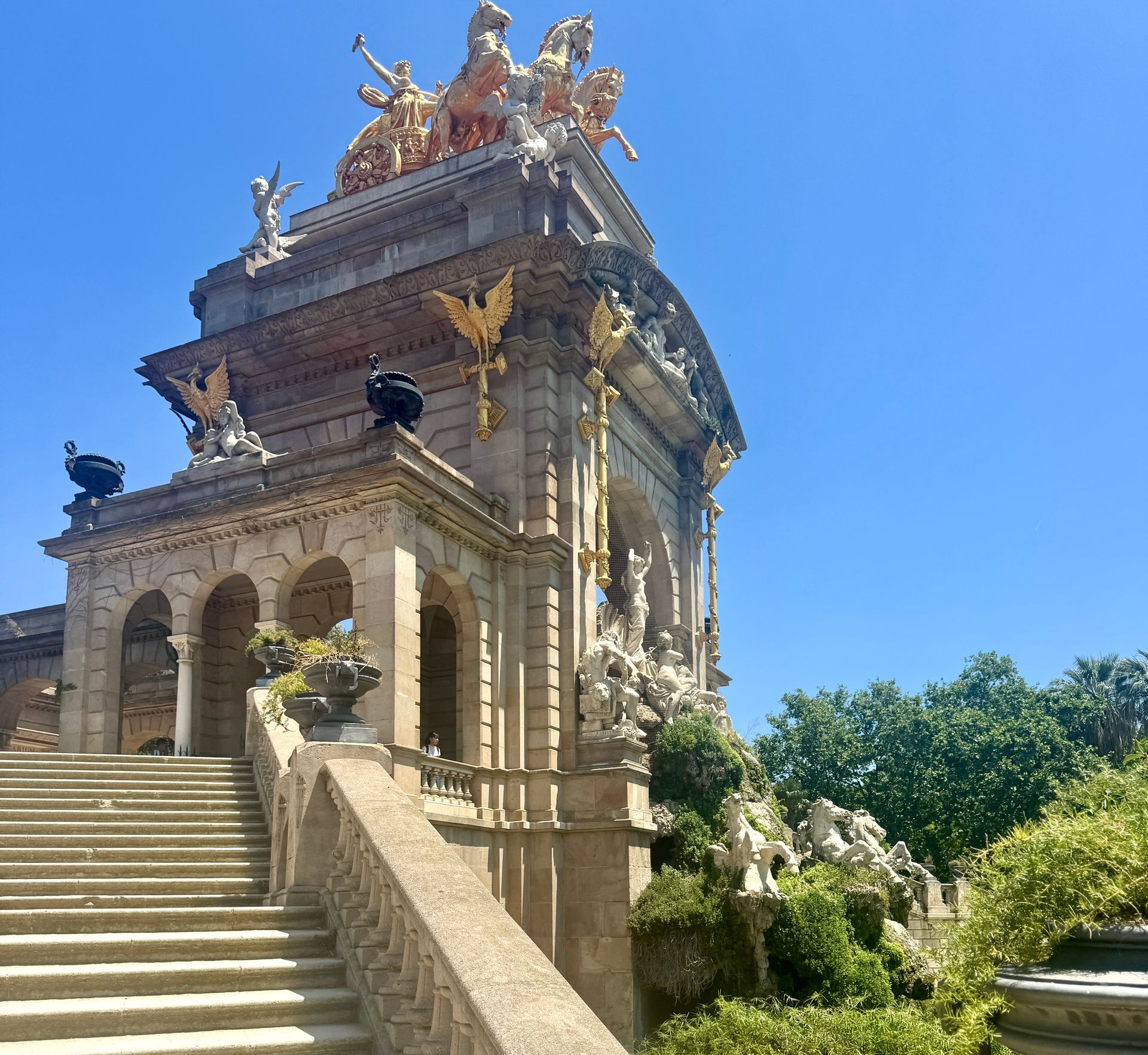 Stone fountain and stairs in a park, topped with gold sculptures against a blue sky.