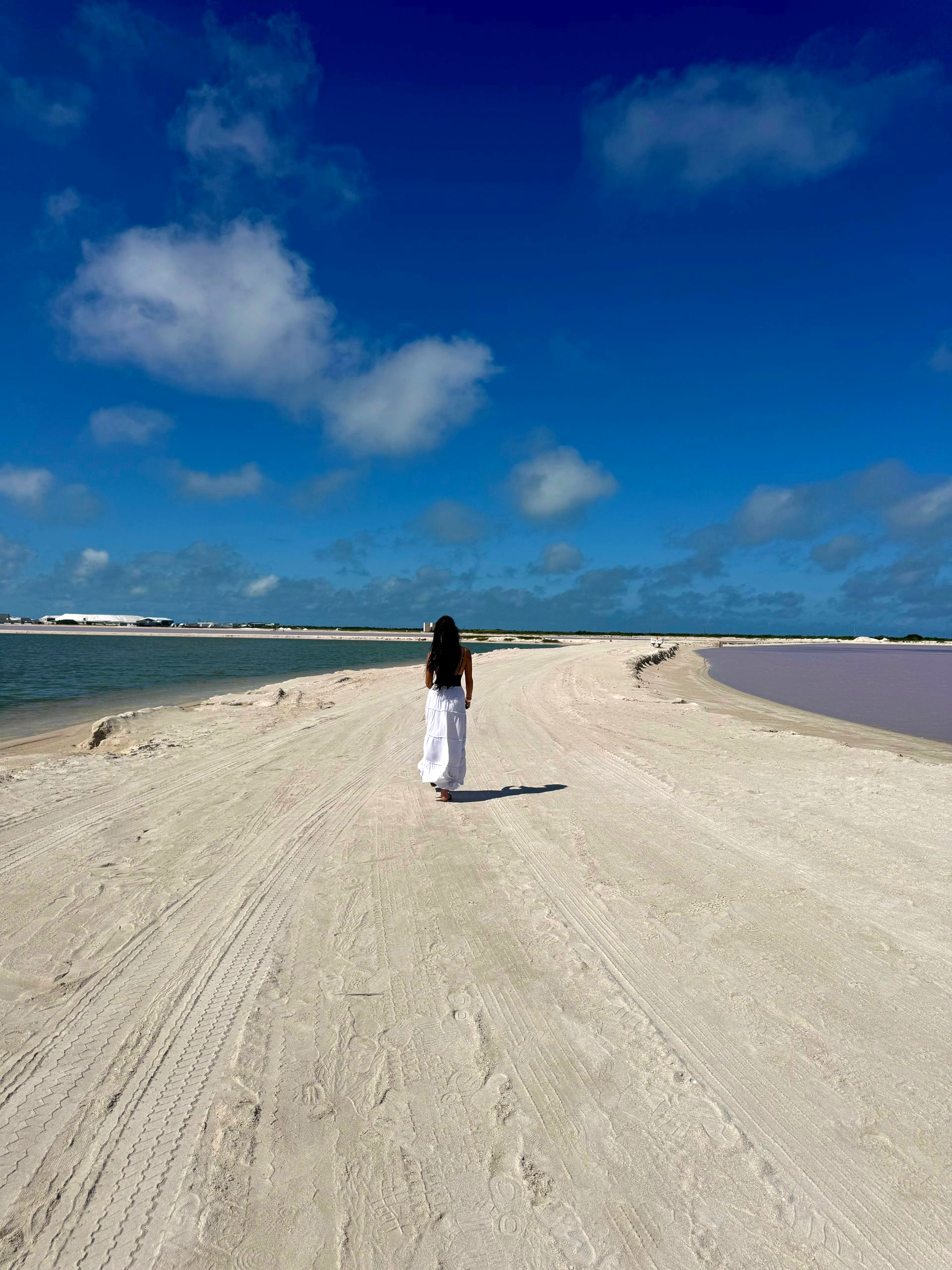 Woman in white dress walks on a sandy path between blue and pink water under a bright, partly cloudy sky.
