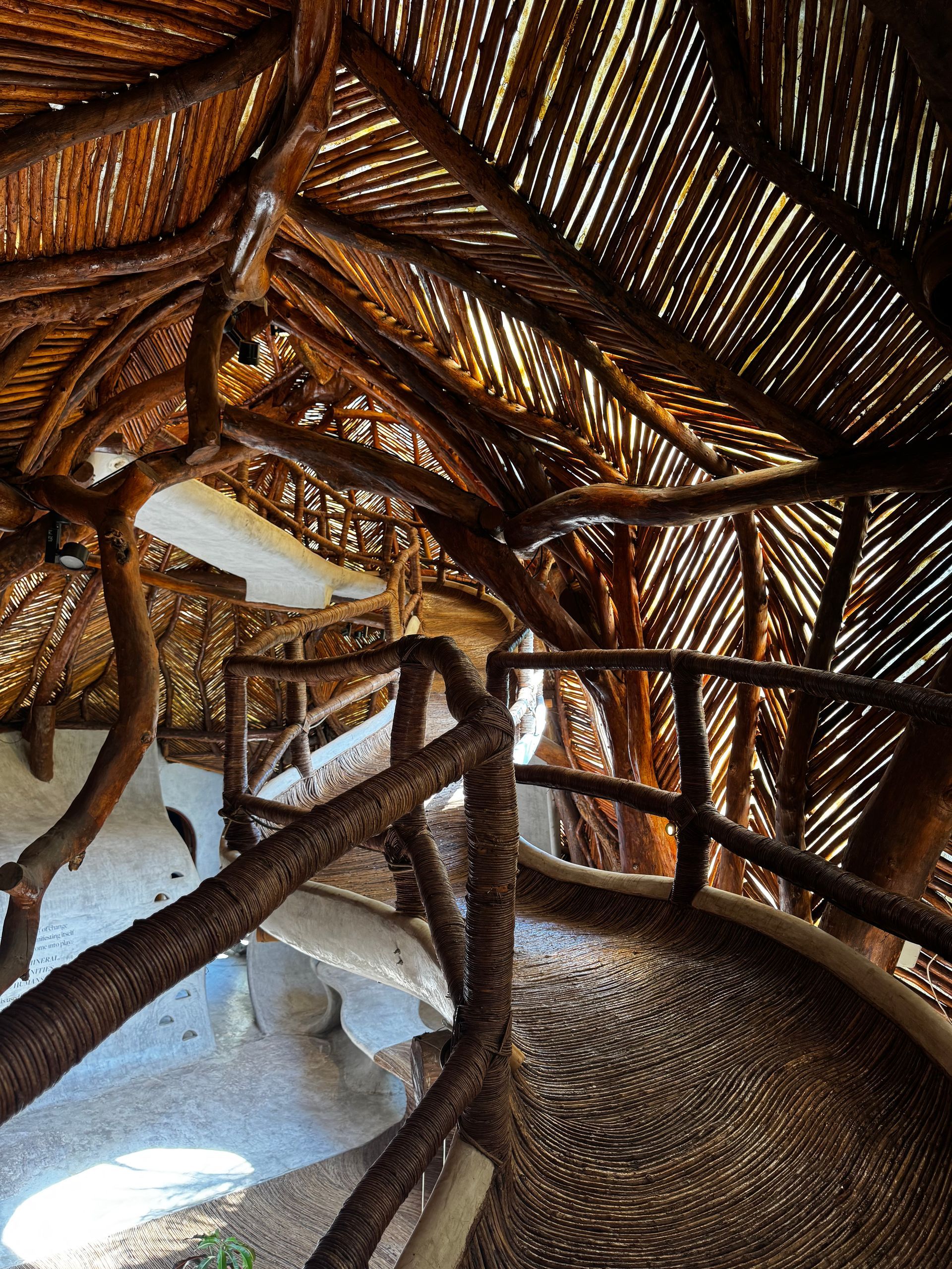 Interior of a rustic building with woven thatched ceiling, wooden support beams, and a curved walkway with railing.