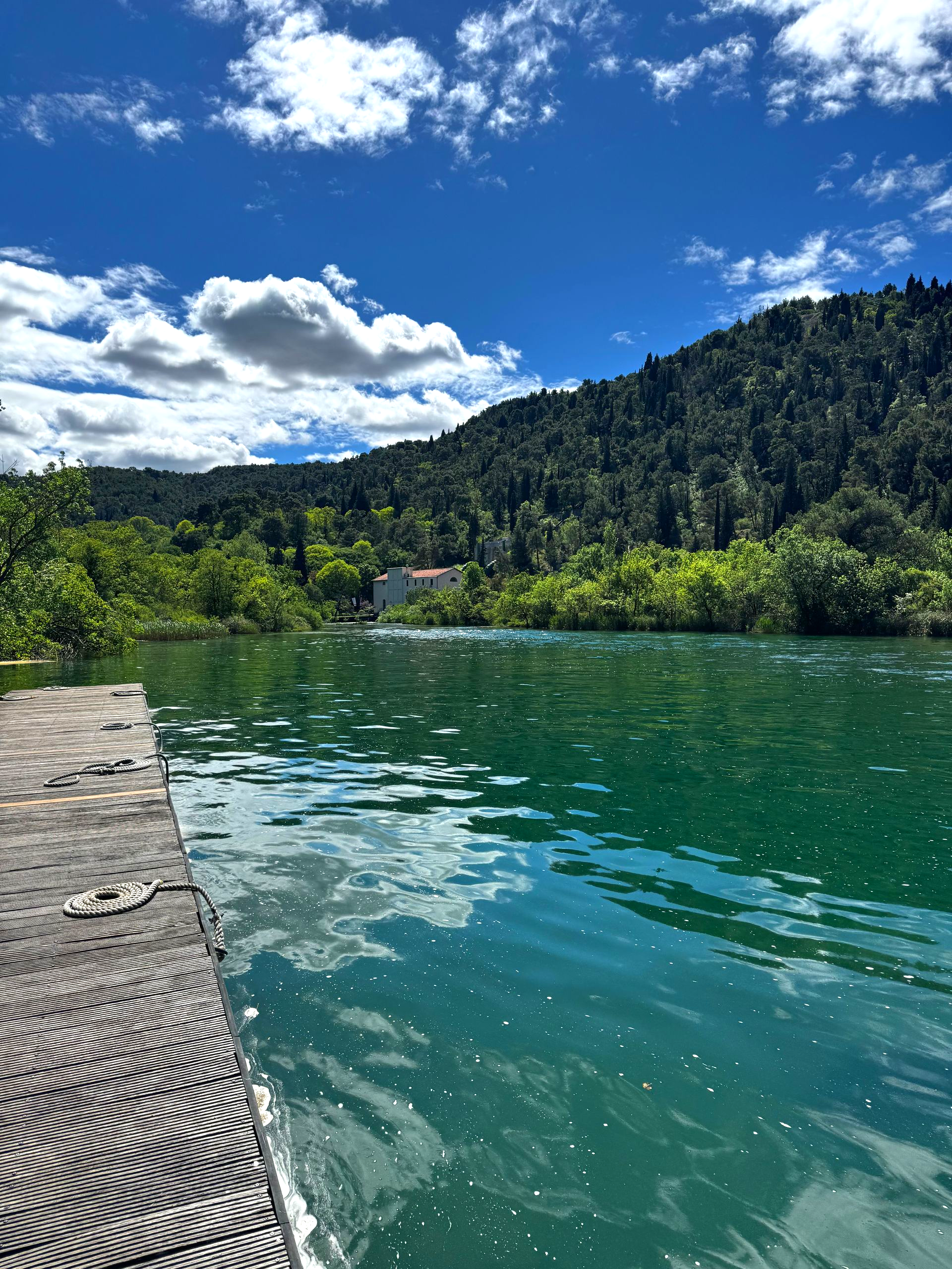 A tranquil turquoise river, edged by a concrete dock, reflects the blue sky with fluffy clouds and lush green hills.