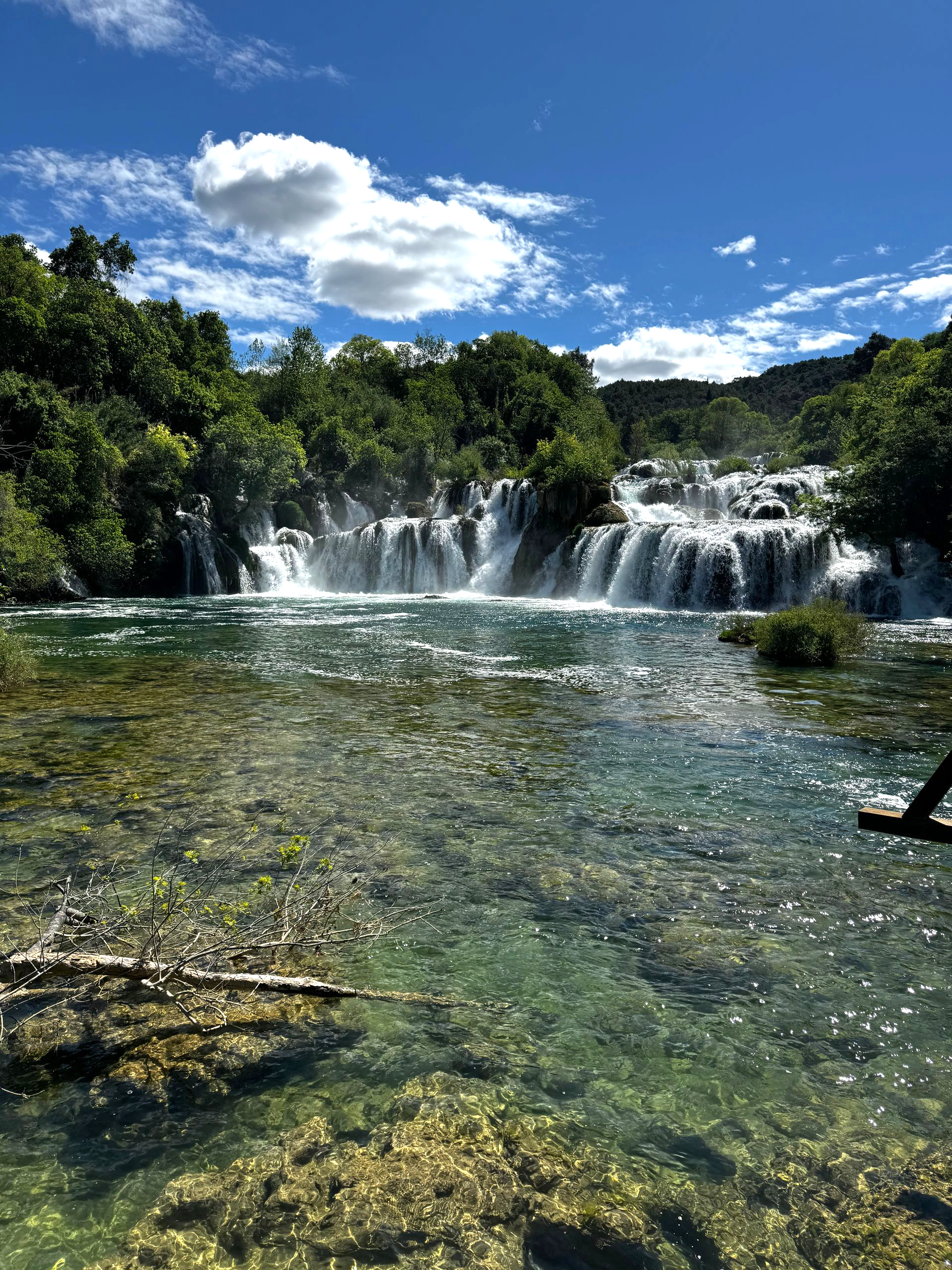 Waterfall cascading into clear water under a blue sky with clouds, surrounded by lush green trees.