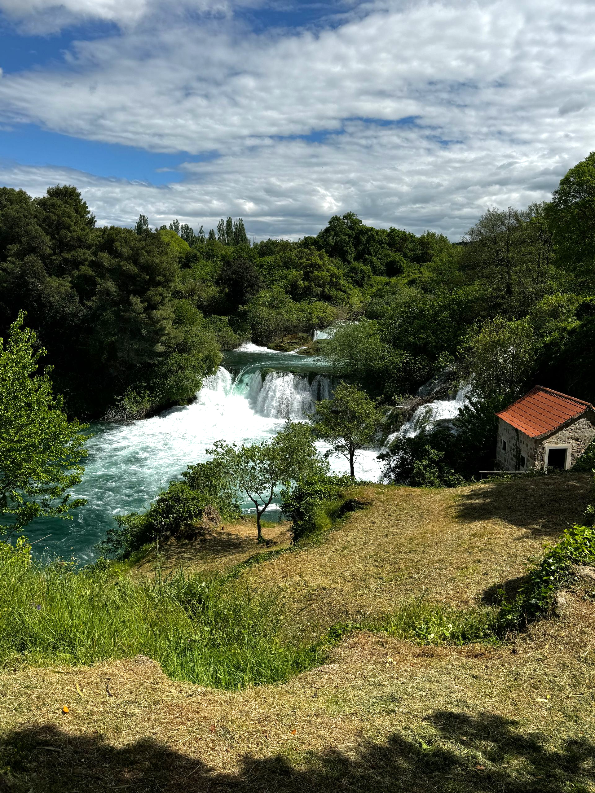 Waterfall cascading into turquoise water, surrounded by lush green trees and a small stone building under a blue sky.