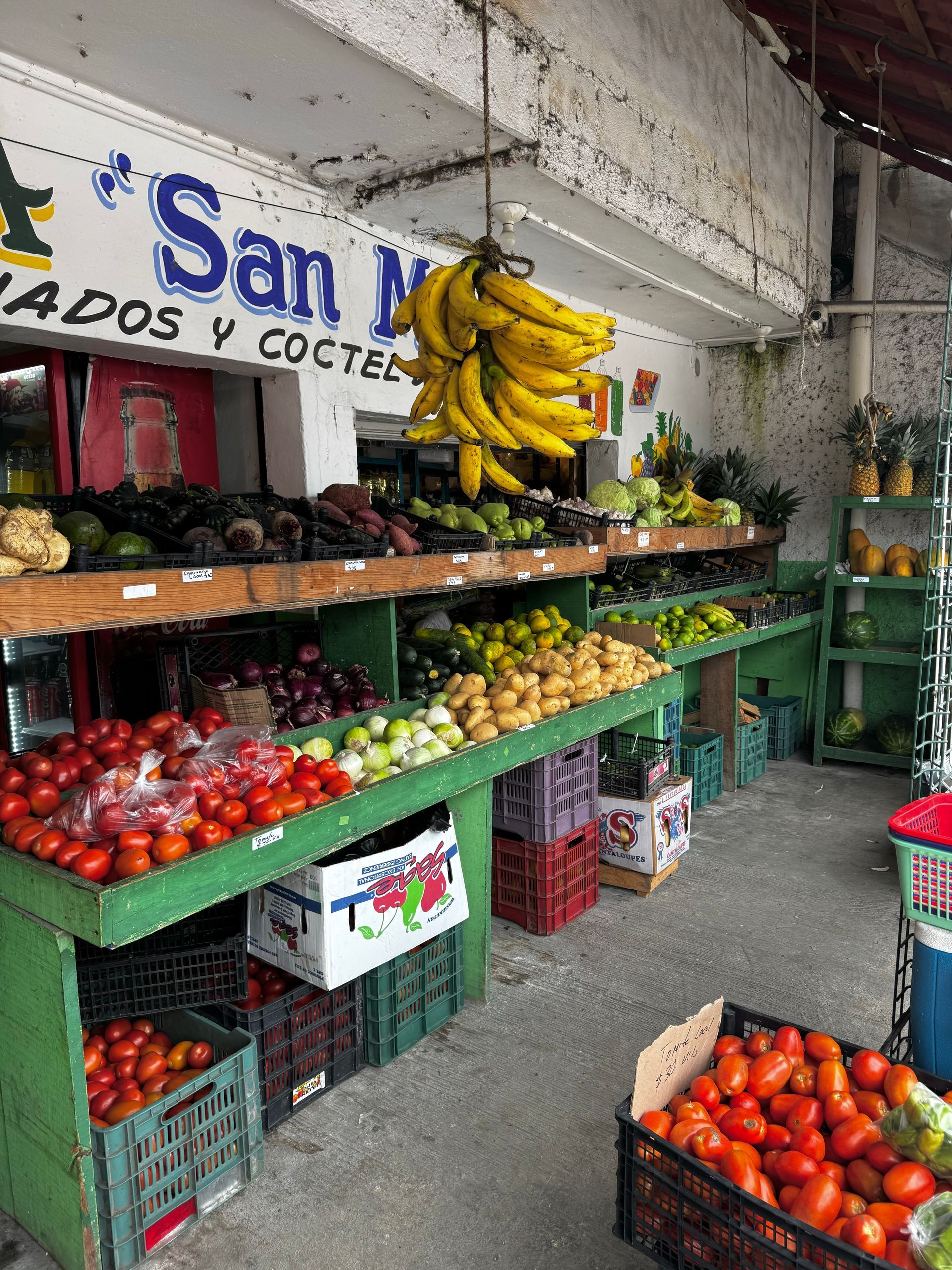 A vibrant fruit stand in front of a shop, showcasing tomatoes, onions, limes, and bananas in Tulum, Mexico.