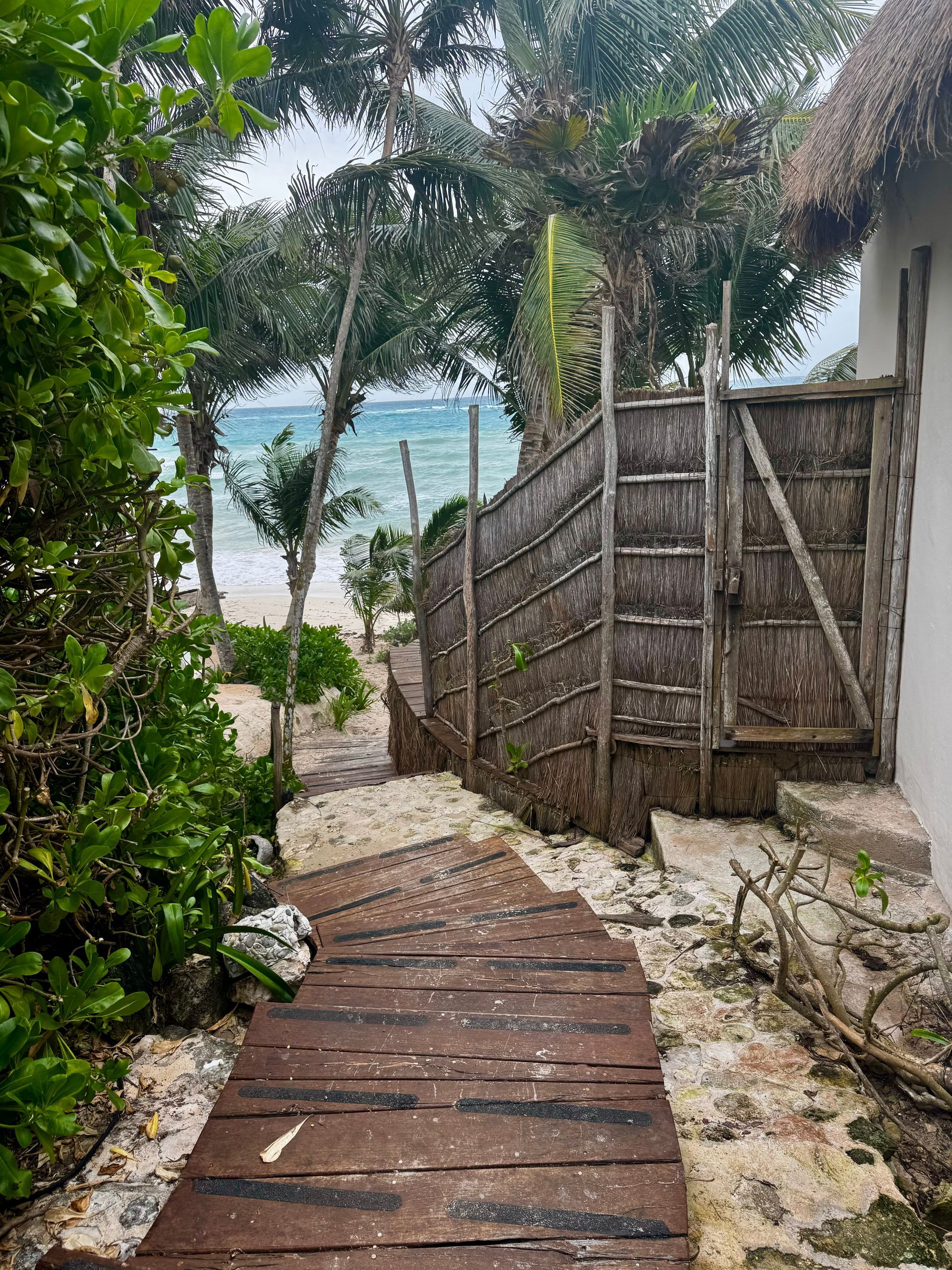 Wooden walkway through tropical foliage leads to a beach, with a gate and a thatched-roof building visible in Tulum, Mexico.