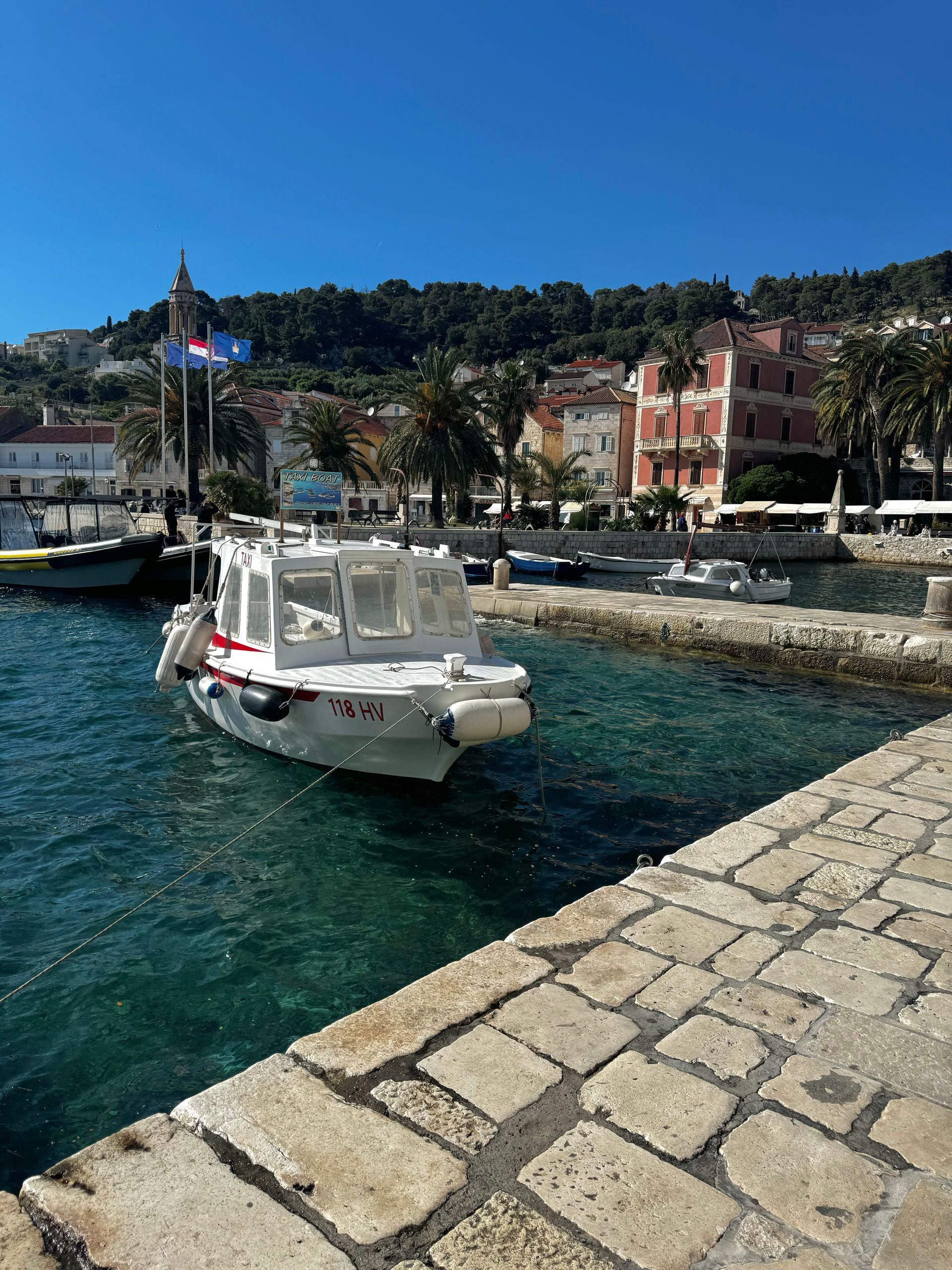 White boat in a harbor with buildings and a hillside in the background, under a blue sky.