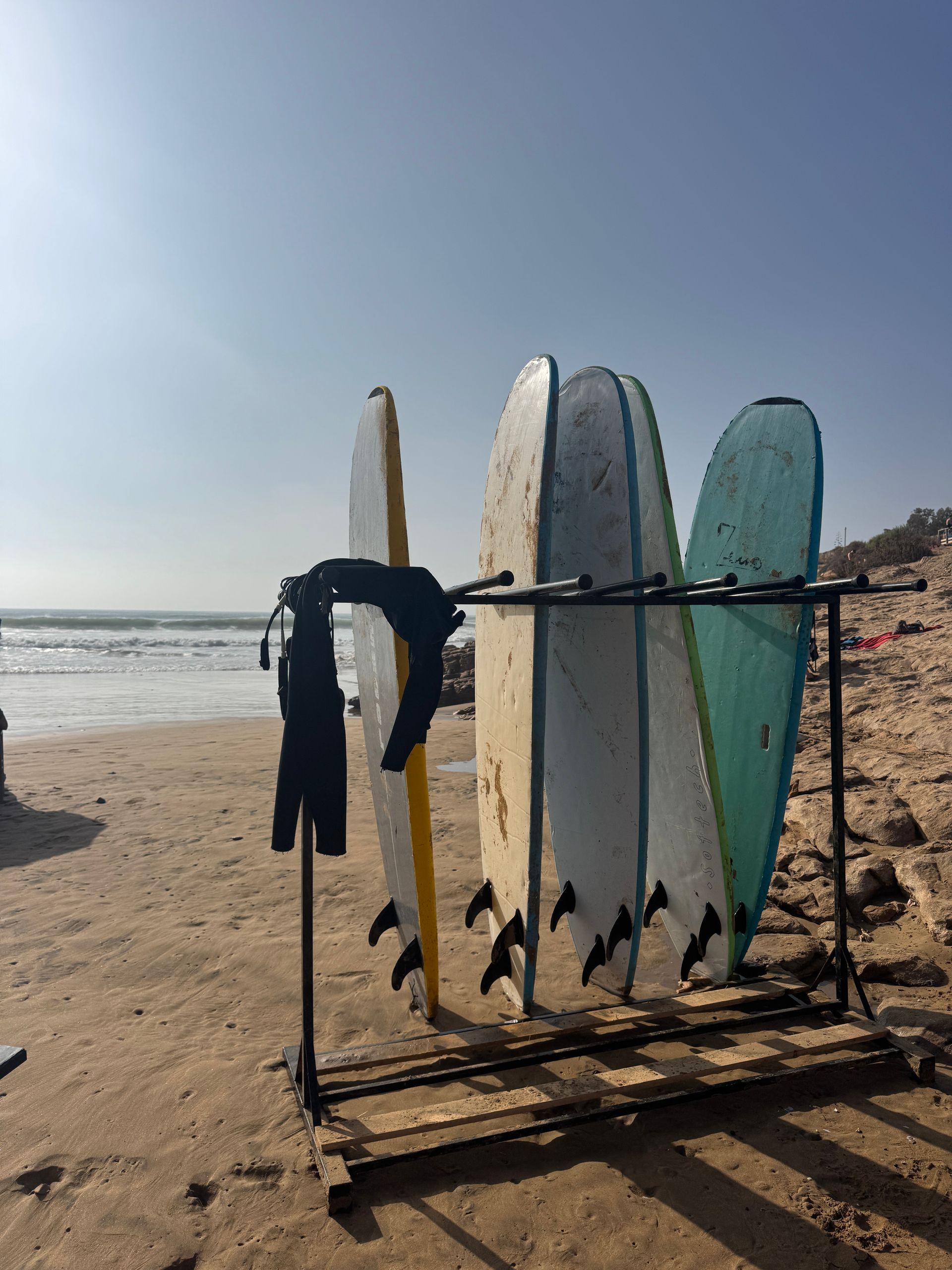 Surfboards on a rack at the beach, with waves in the background, a wetsuit hanging.