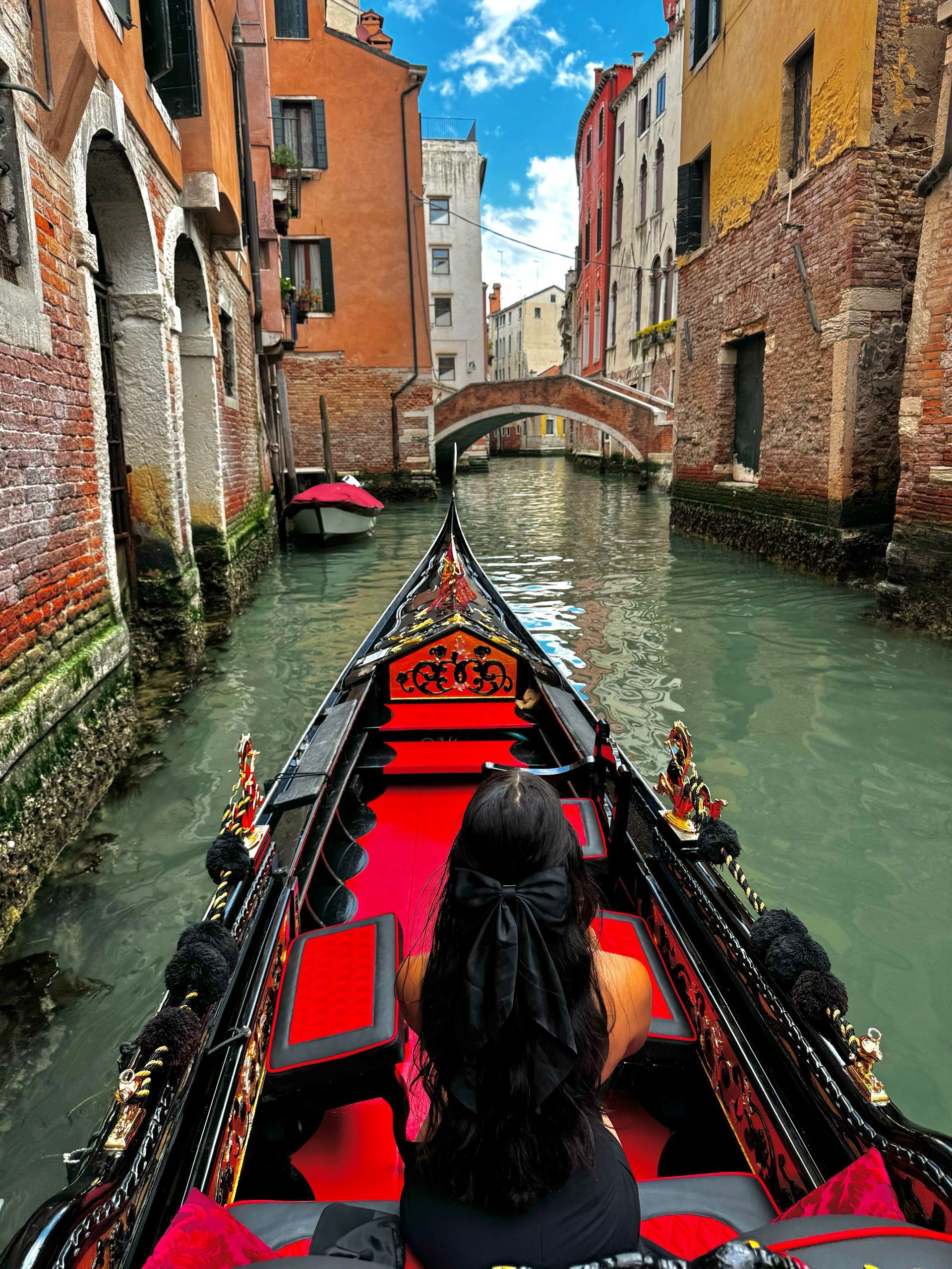 Woman on a gondola in Venice canal, looking towards bridge and colorful buildings.