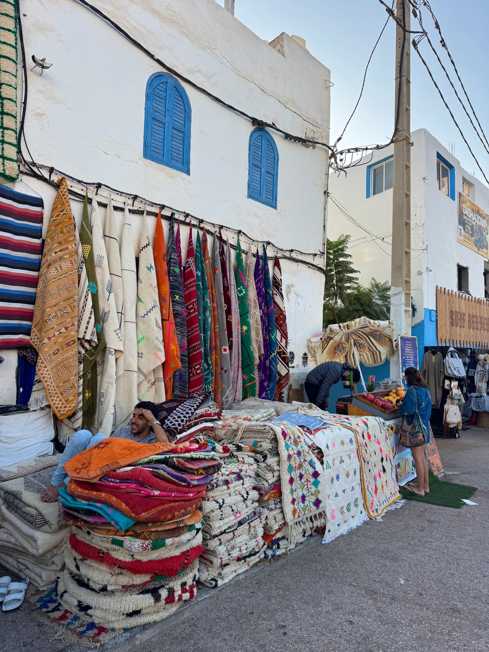 Shopfront displaying colorful rugs in a white-walled building with blue shutters. People browse goods on cobblestone street.