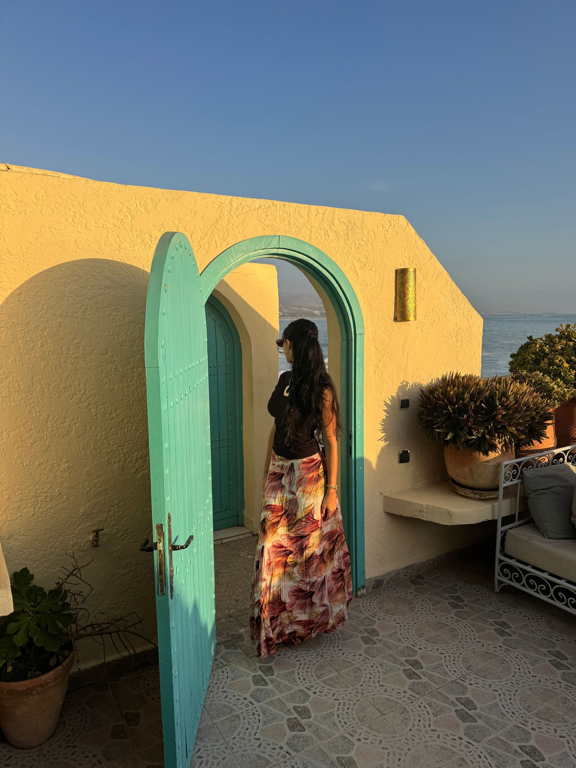 Woman in floral skirt stands in a turquoise doorway, overlooking the ocean. The building is beige with potted plants.