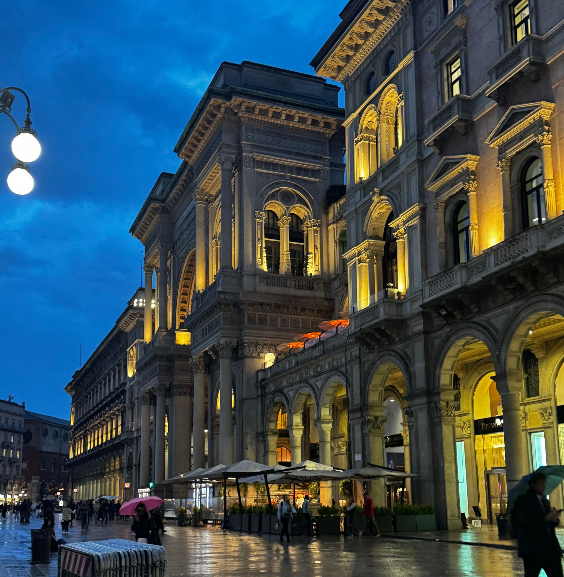 Elegant European street lit at night. Rain-slicked pavement reflects golden light from buildings, diners, and streetlights.