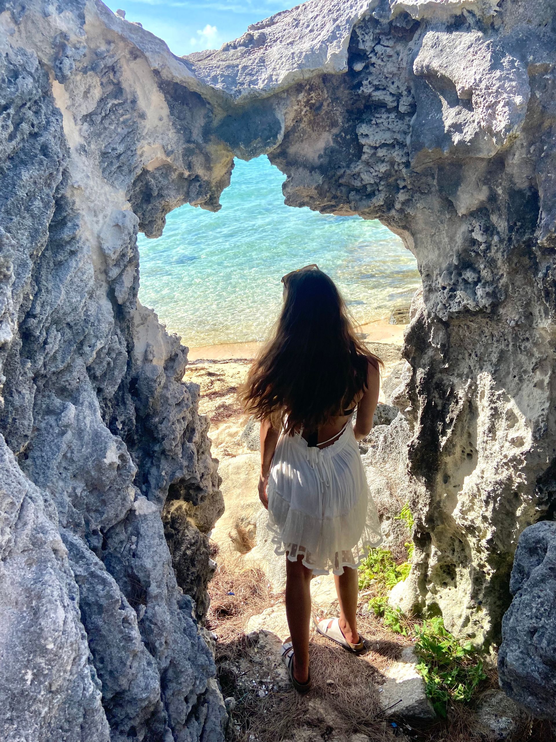 Woman in white dress looks through a rocky archway at a turquoise ocean.