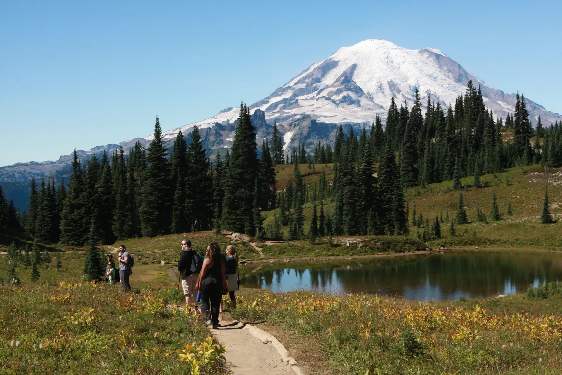 A group of people are walking along a path next to a lake with a mountain in the background in Mount Rainier.