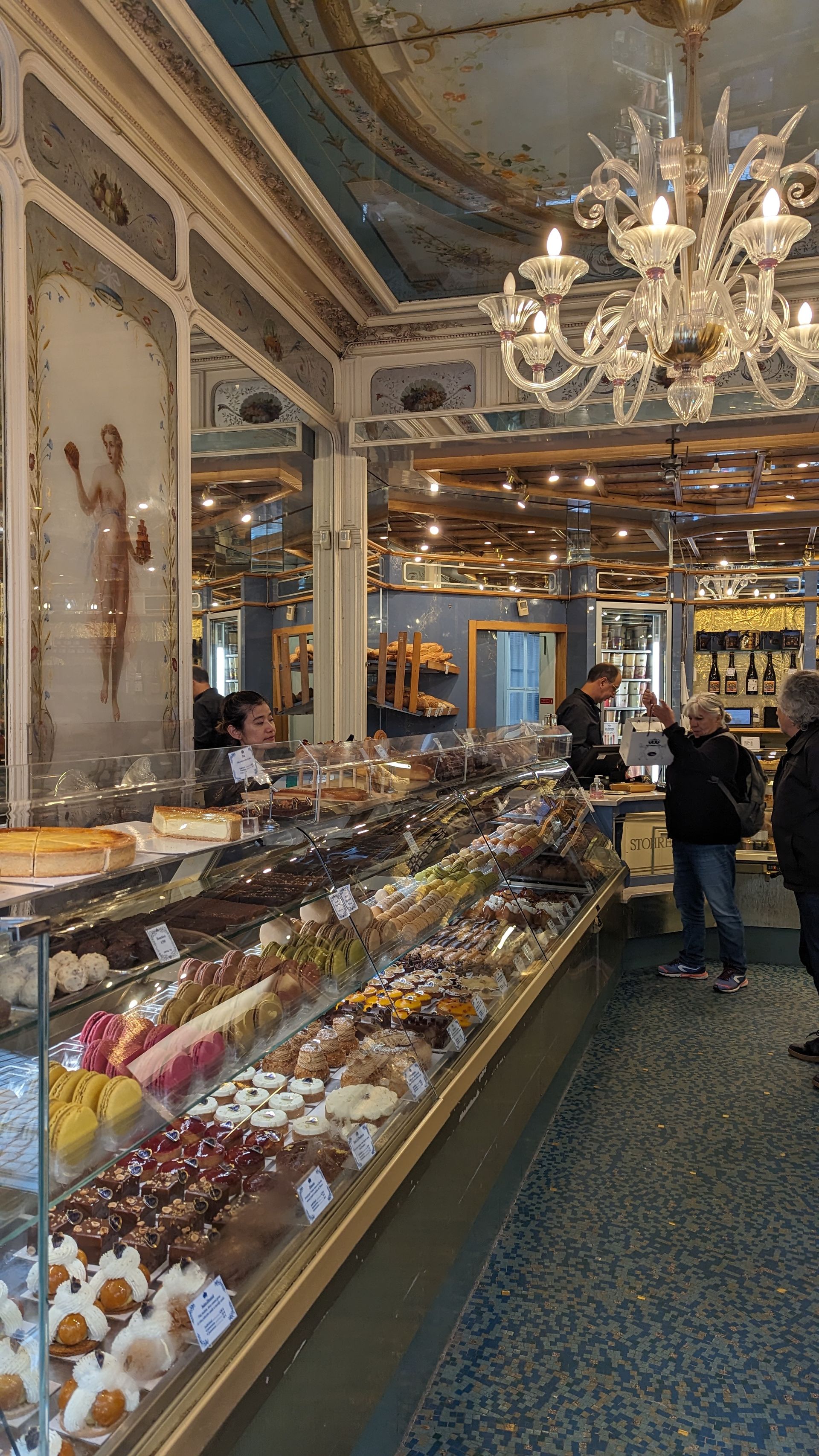 Inside a pastry shop: display cases full of treats, customers and staff. Ornate ceiling, chandelier.