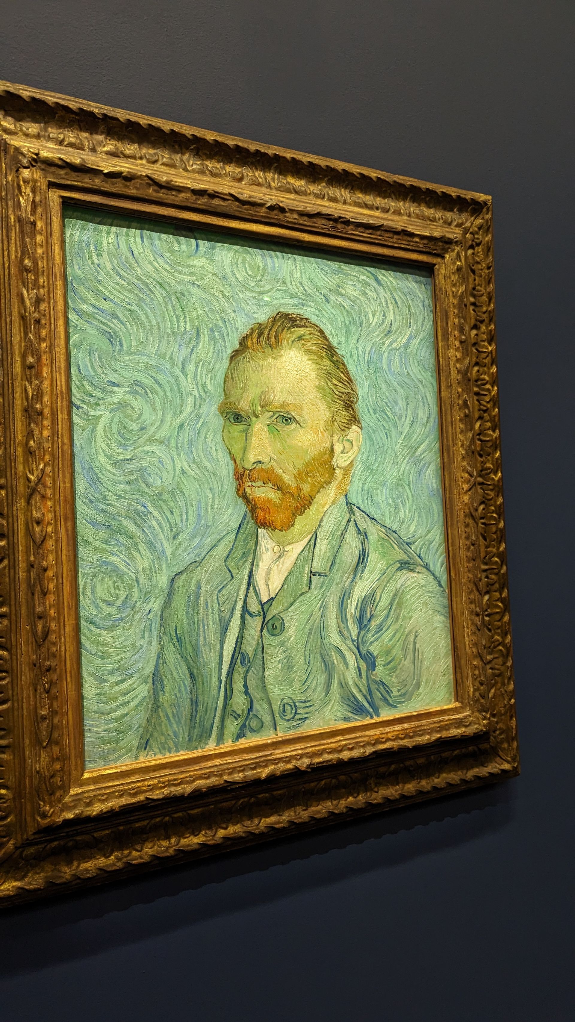 Van Gogh's self-portrait in ornate golden frame, on blue wall. Man with orange beard, green jacket, gazing forward.