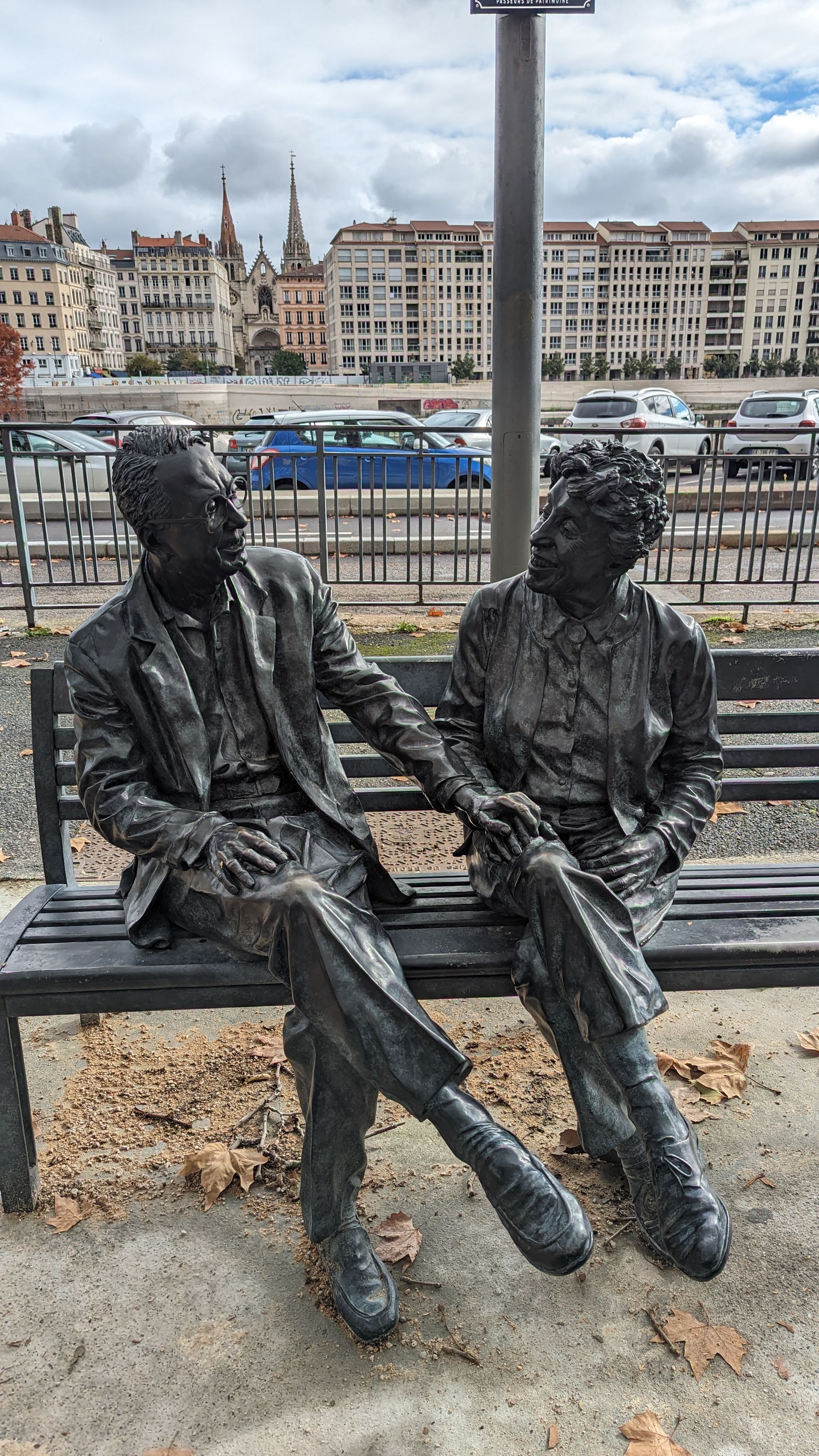 Bronze statue of a couple sitting on a bench, holding hands. City buildings in the background.