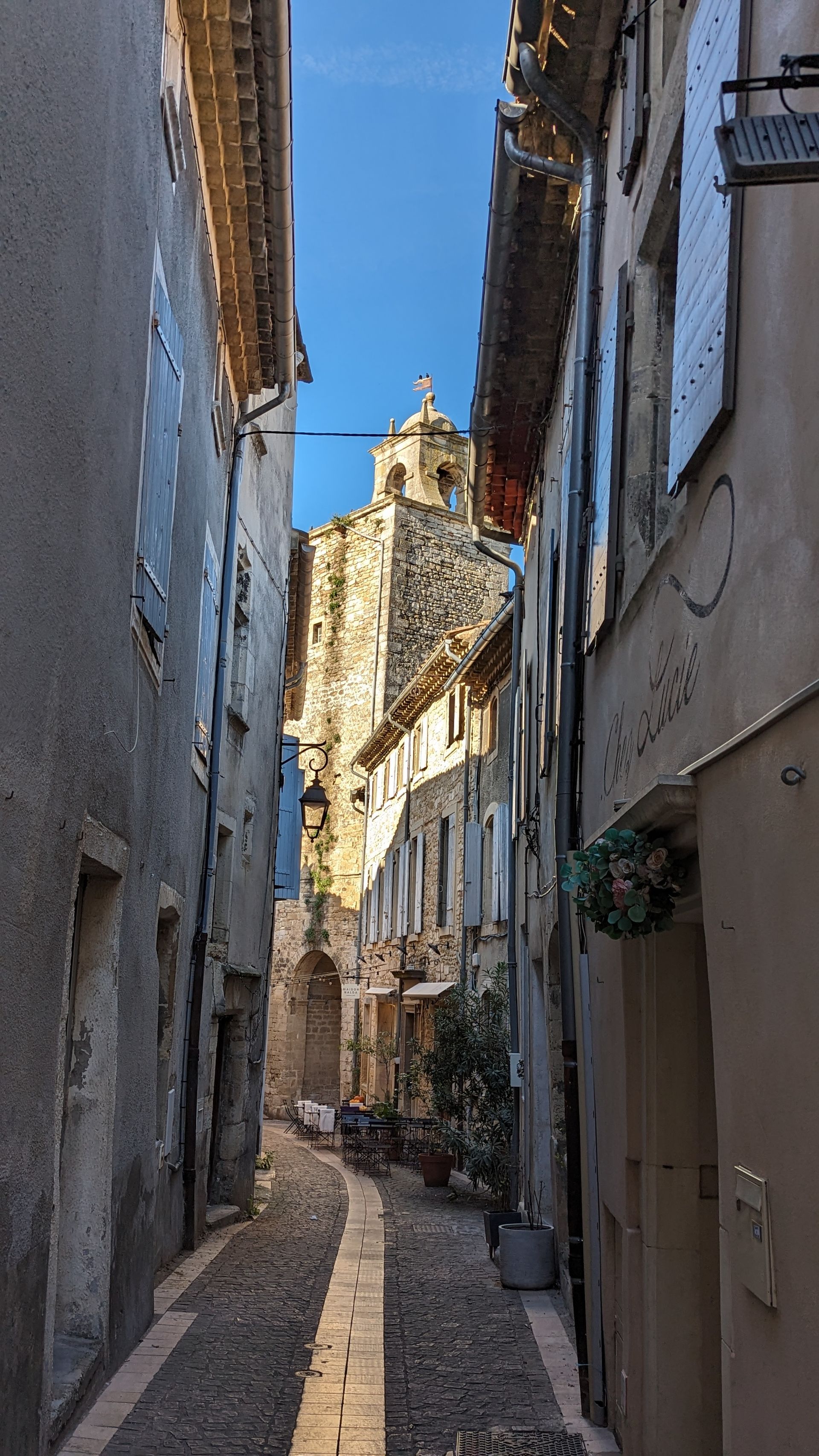 Narrow cobblestone street in a European town, with tall buildings and church steeple visible. Blue sky.