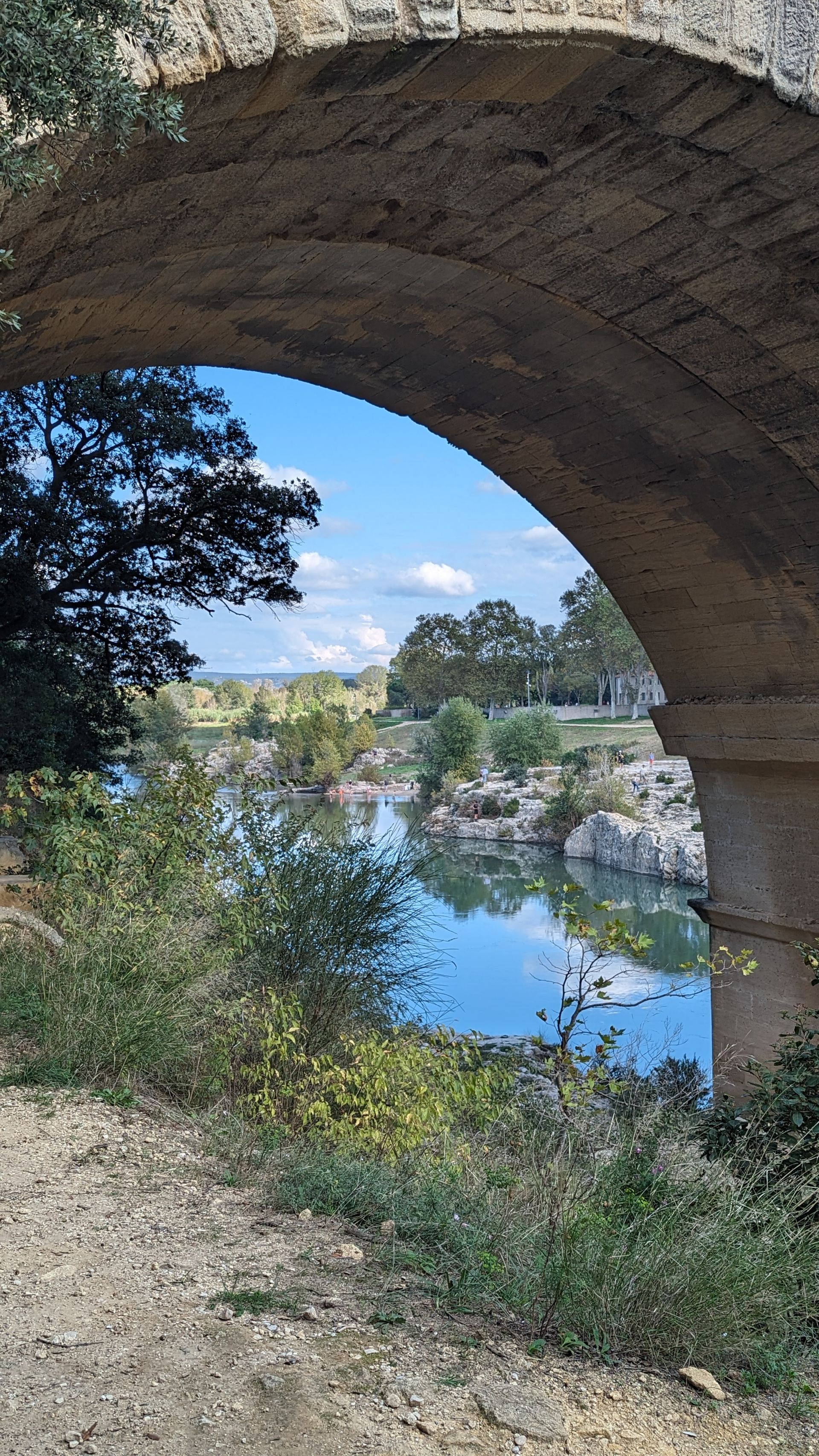 View through an arch of an old stone bridge, showing a river, blue sky, and surrounding trees.