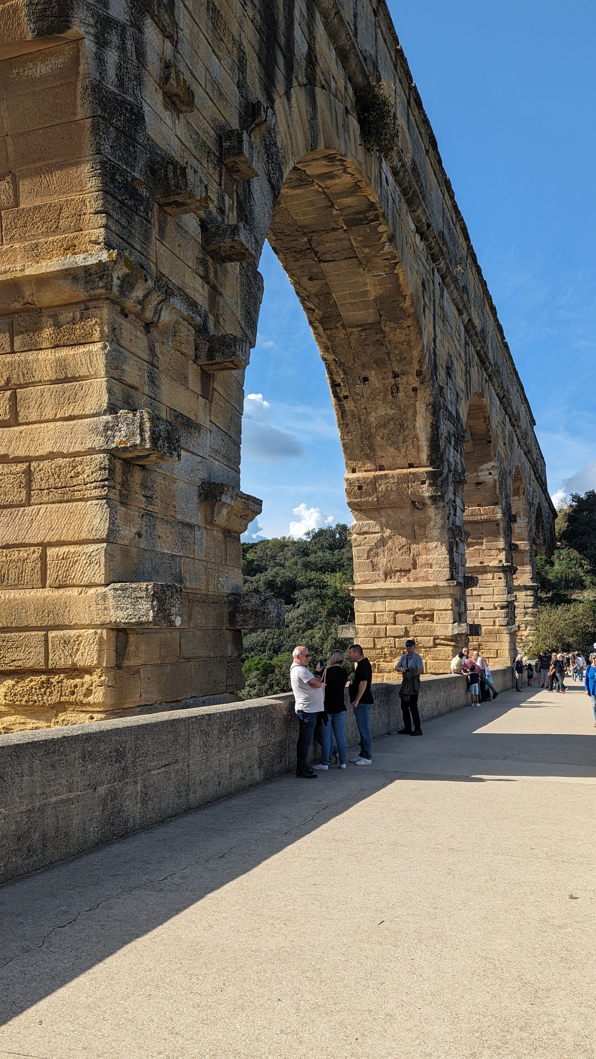 Ancient stone aqueduct with people standing on a pathway below the arches, under a blue sky.