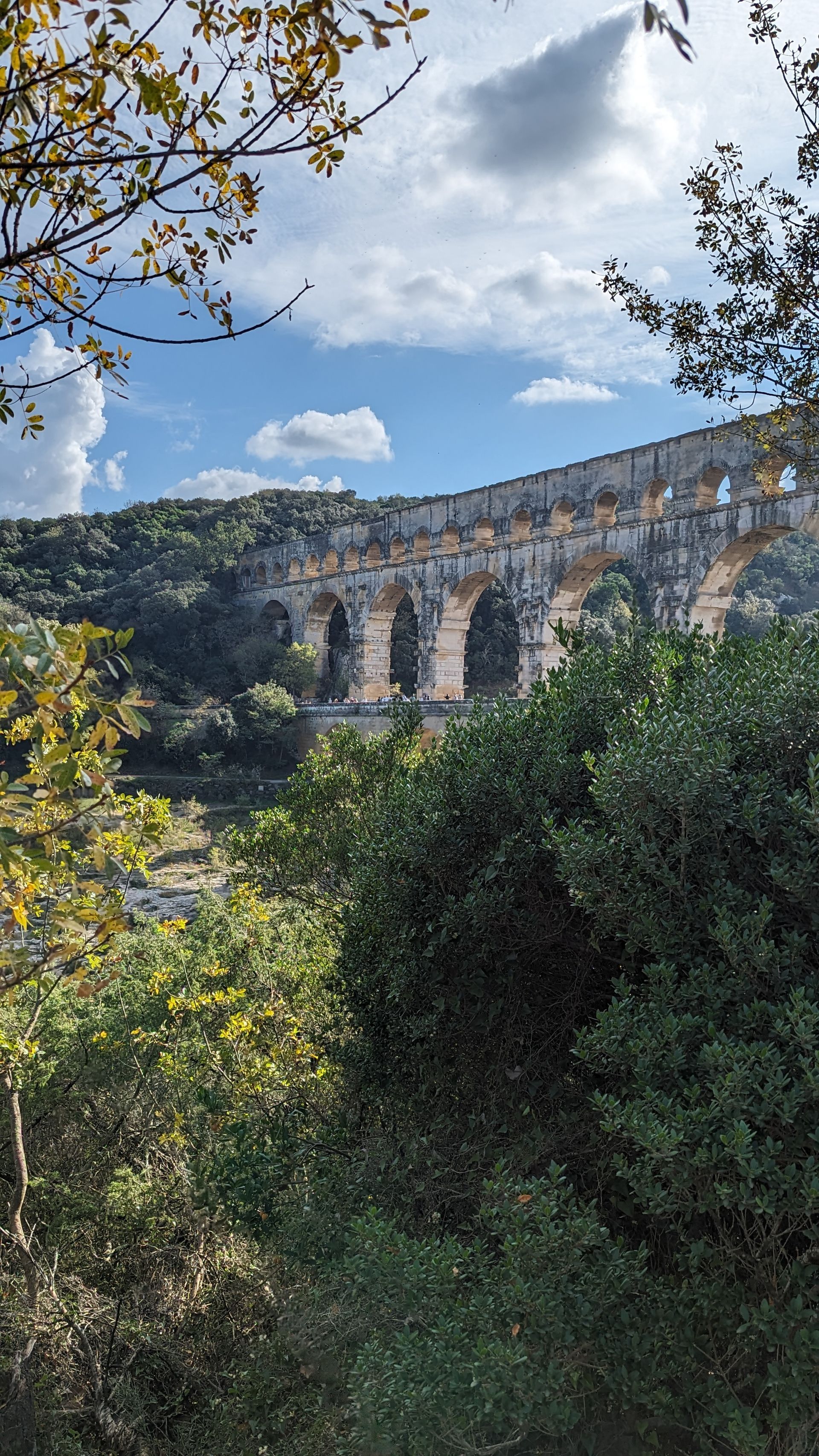 Roman aqueduct arches against a backdrop of trees and a partly cloudy blue sky.