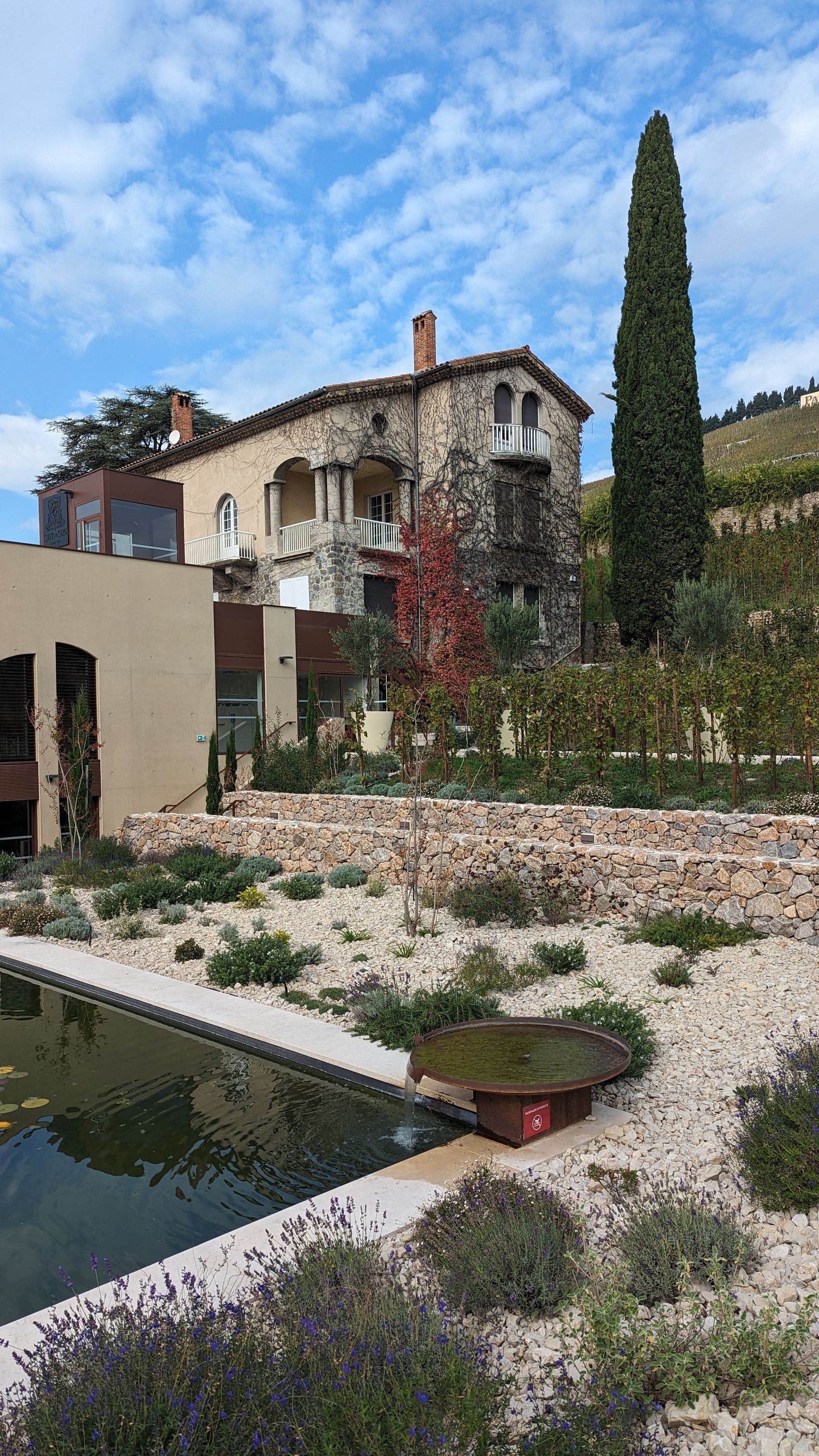 Stone building with balcony, tall tree, and pond in a garden setting on a hillside under a cloudy sky.