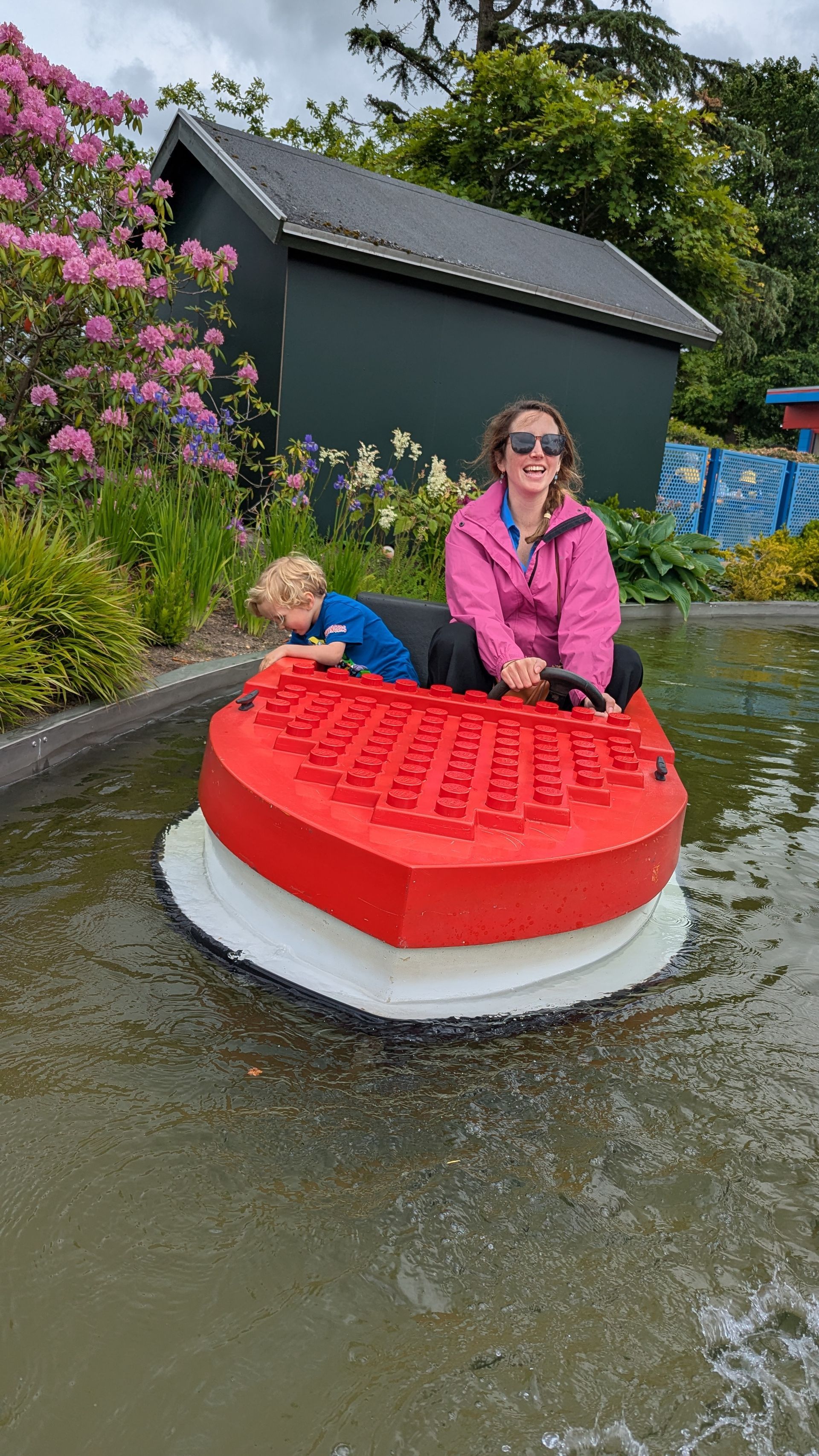 Woman and child in a red Lego boat on a pond; smiling, with building and flowers in background.