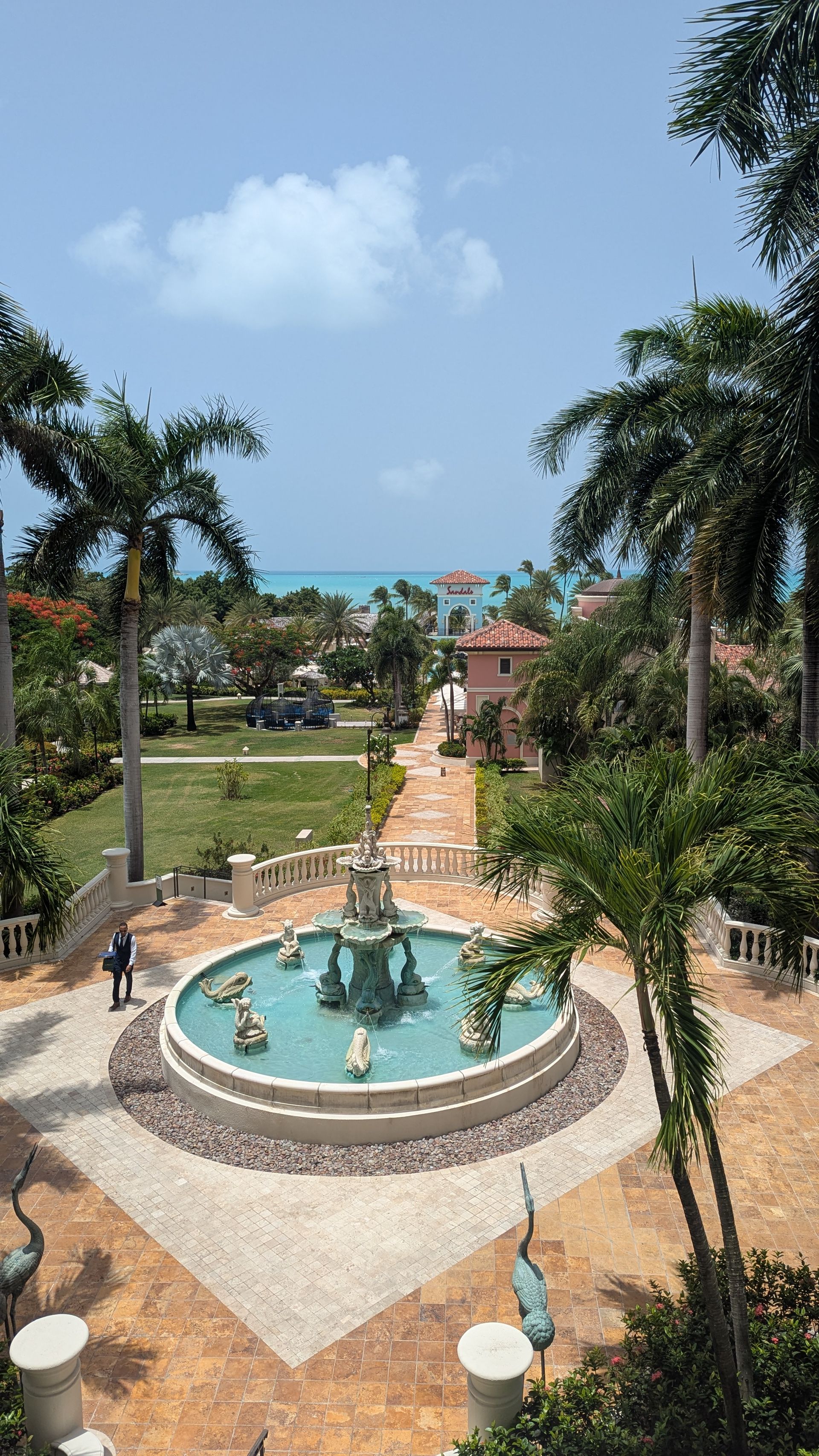 Fountain in a courtyard with a pathway leading to the ocean; palm trees, blue sky.
