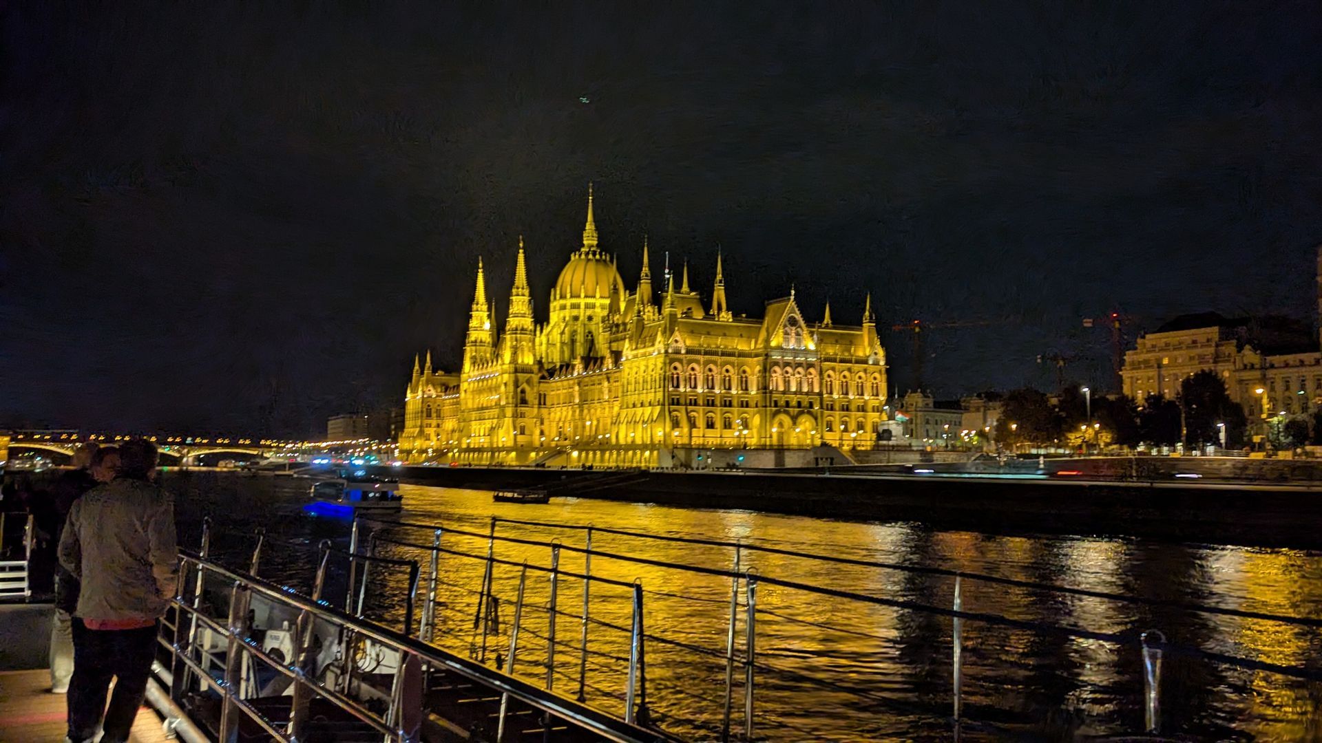 Hungarian Parliament Building illuminated at night, reflected in the Danube River. Person on a boat, distant cityscape.