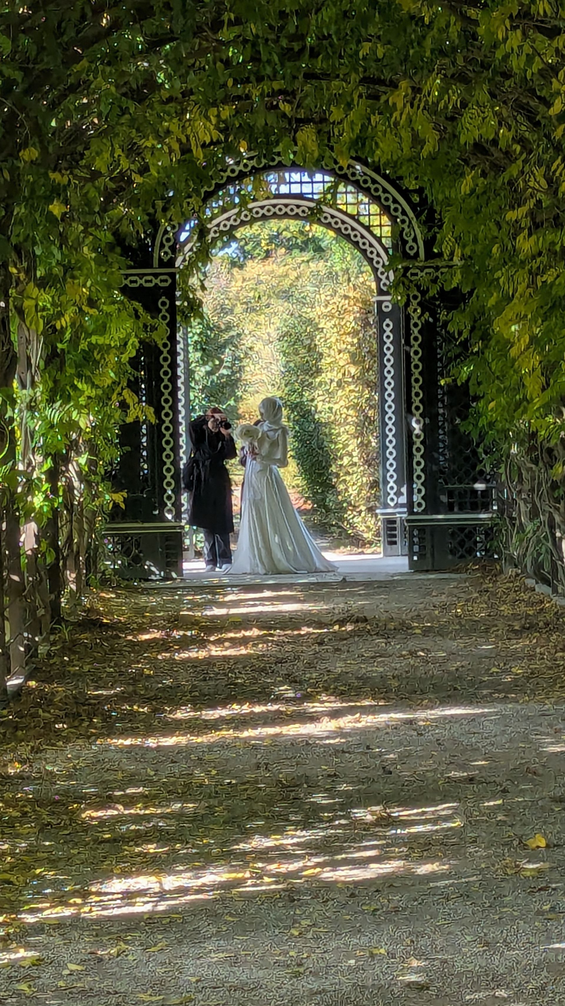 Bride and photographer under an ornate archway, in a garden with autumn foliage.