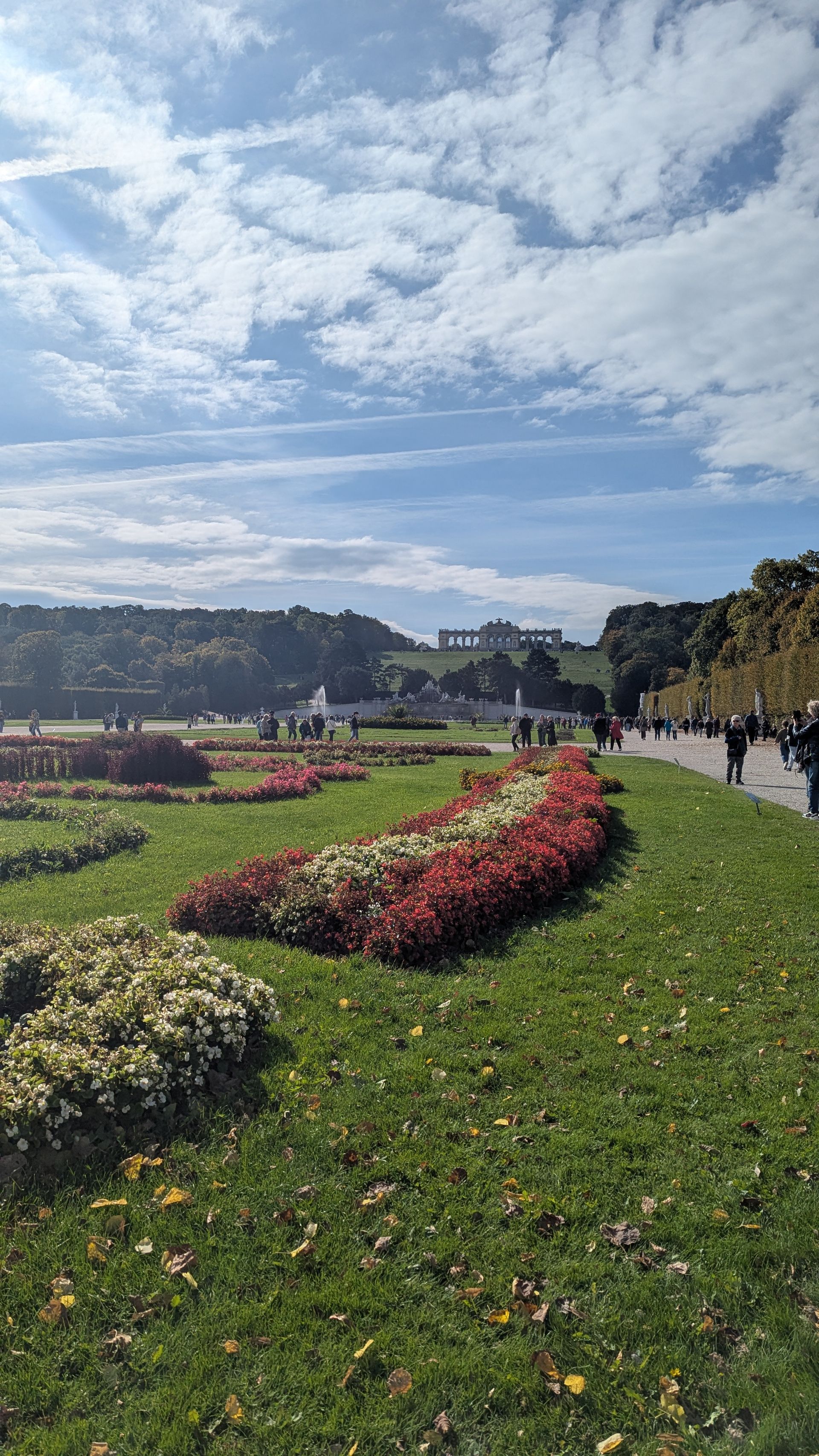 Flower beds in a park under a cloudy blue sky. People walk a pathway.